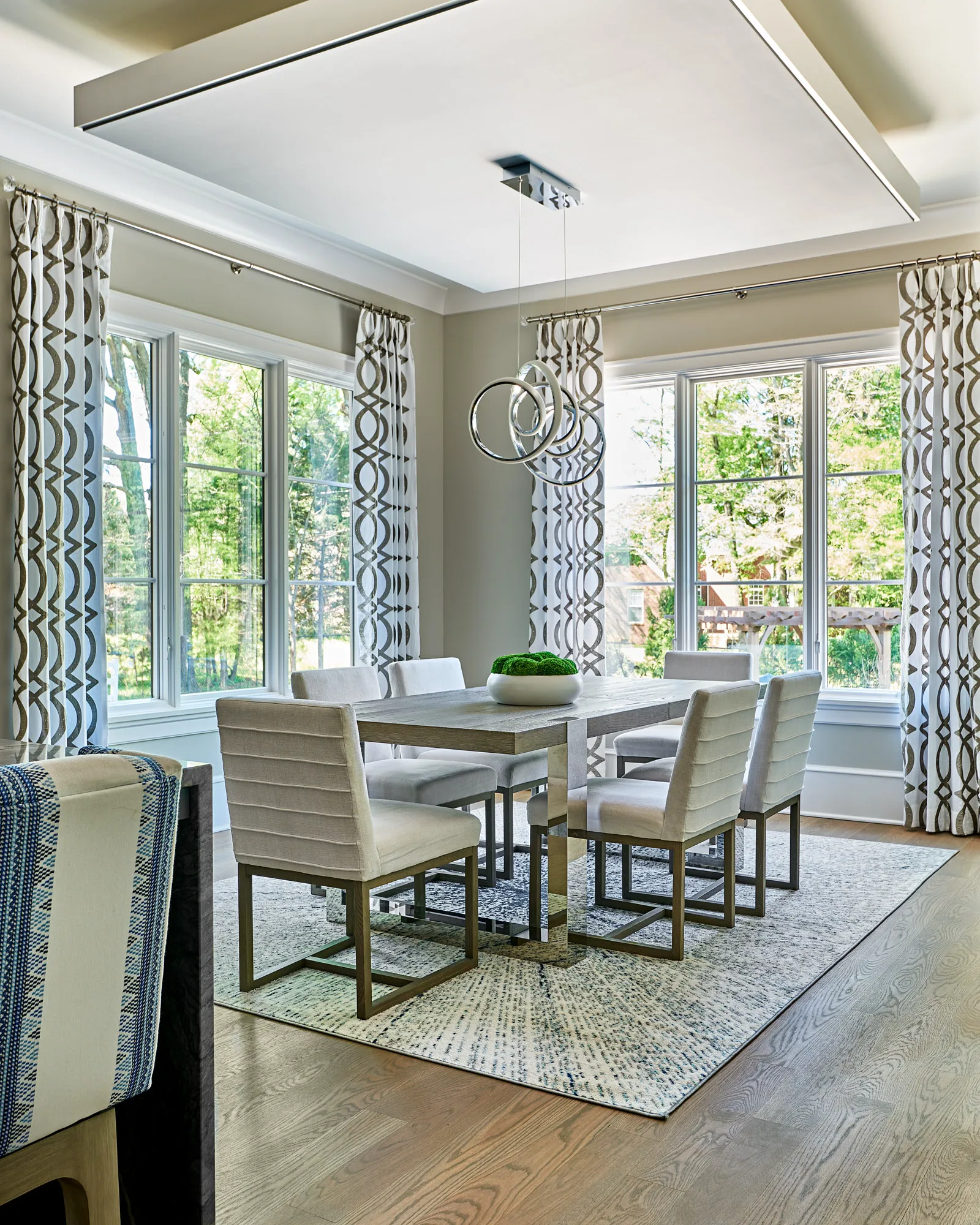 A bright, modern dining room featuring a grey wooden table, light upholstered chairs, and a chrome ring chandelier. Geometric curtains frame large windows showing trees.