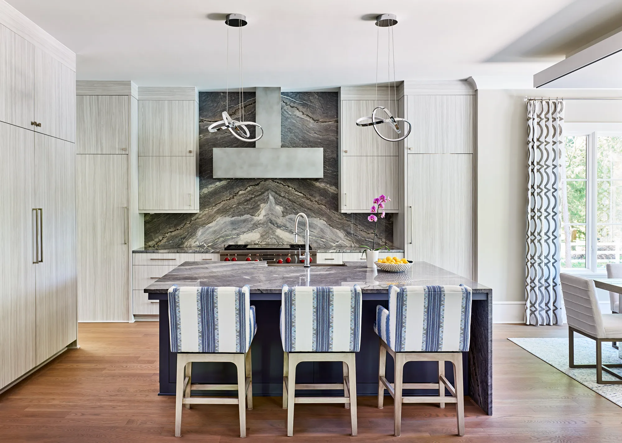 A sleek, modern kitchen features light wood cabinets surrounding a dramatic, book-matched dark stone backsplash. Three blue and white striped barstools sit at the large kitchen island.