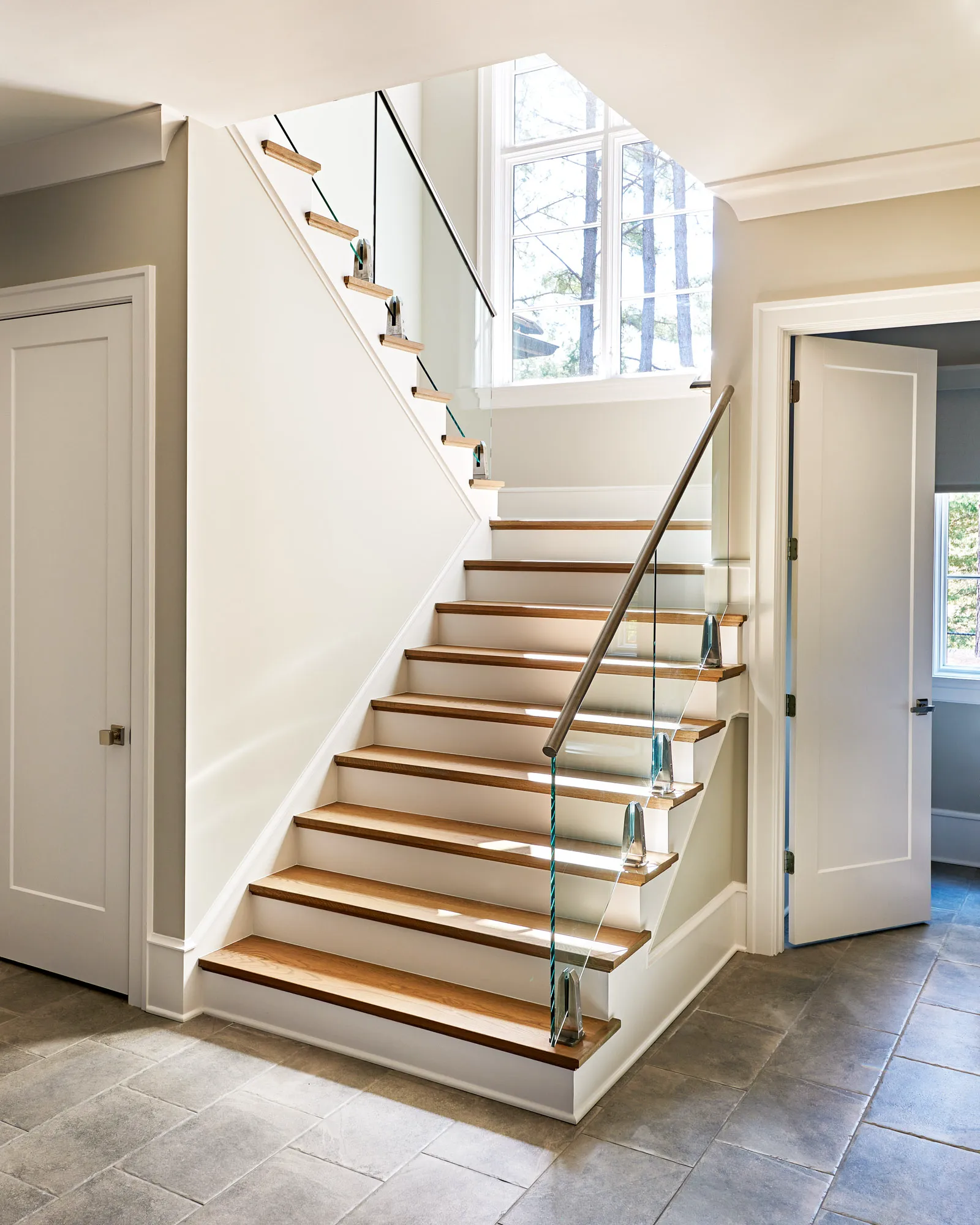 A brightly lit, modern interior staircase features floating wood treads, white risers, and a sleek glass railing with a metal handrail. The entryway has gray stone tile flooring.