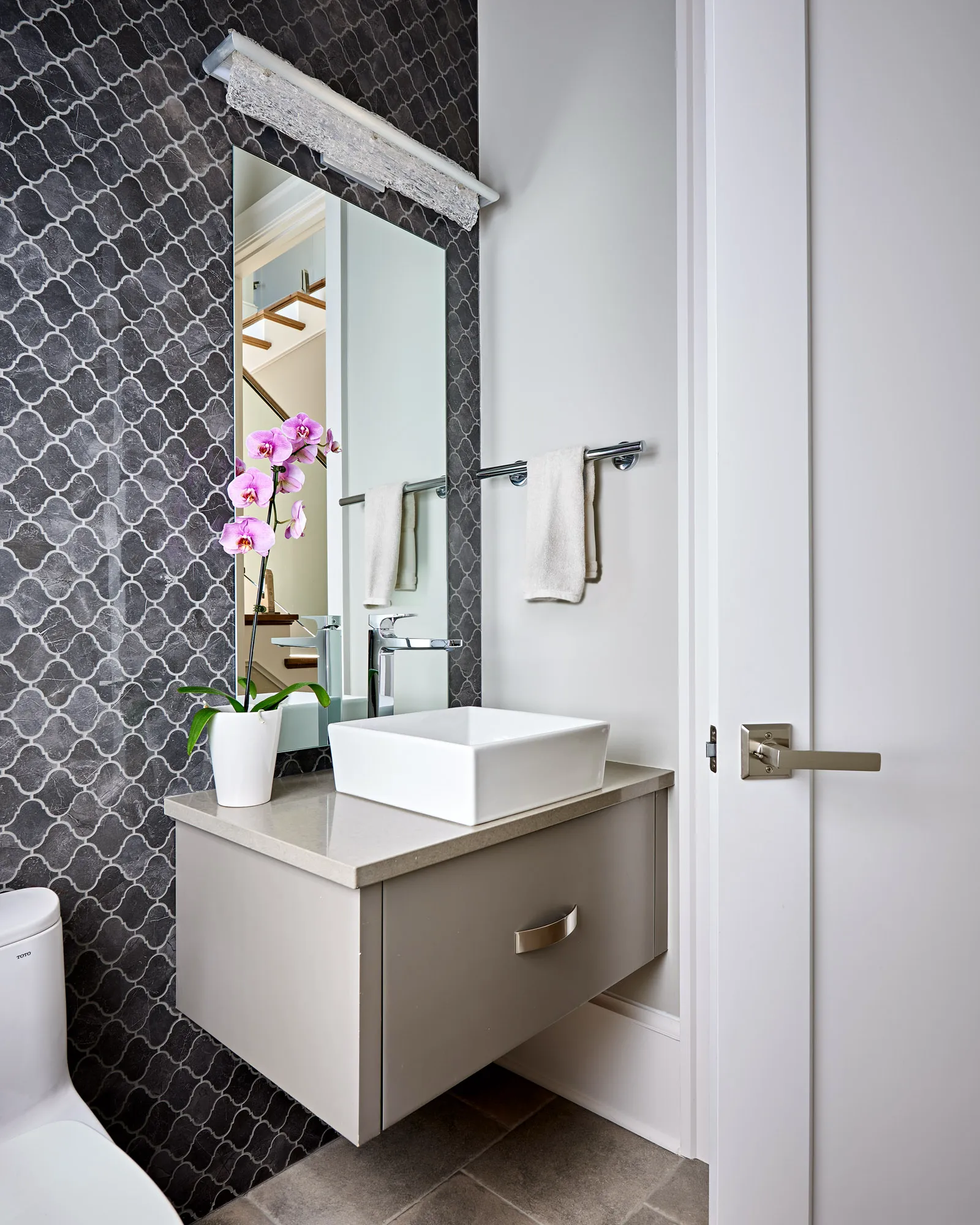 A contemporary powder room with a floating vanity, vessel sink, and pink orchid. An accent wall features dark gray arabesque tile and a textured light fixture.