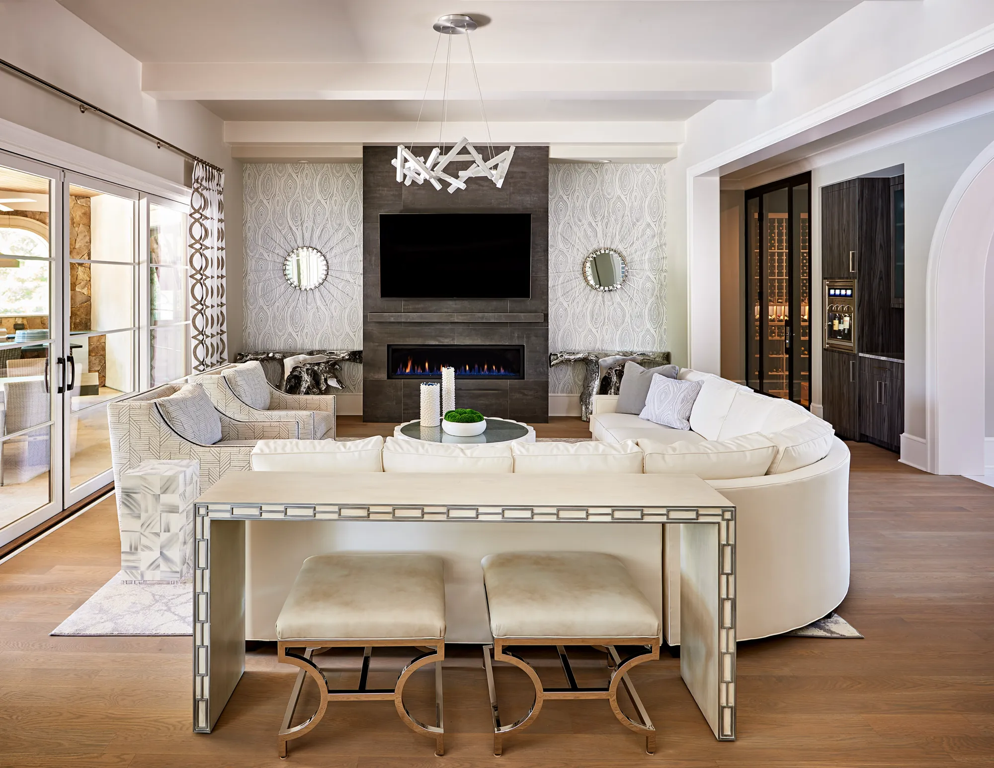 A neutral, contemporary living room with a curved white sectional and chairs facing a dark gray tiled fireplace and television feature wall, with an adjacent wine cellar.