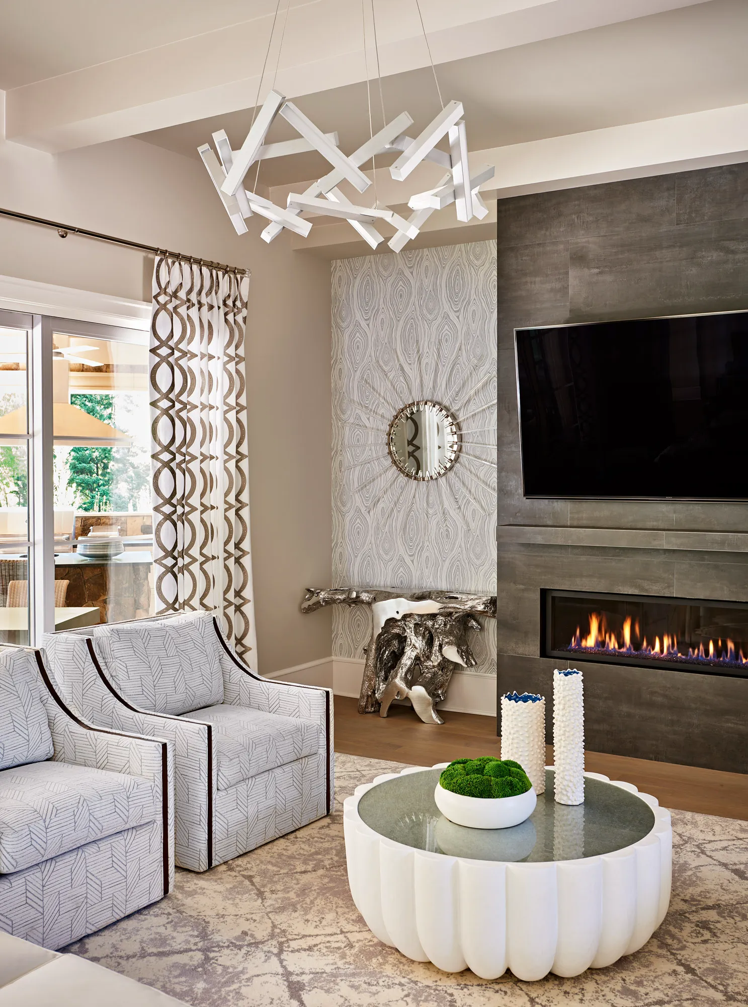 A contemporary living room features patterned armchairs, a rounded coffee table, and a linear fireplace set in a dark tile wall below a striking, angular light fixture.