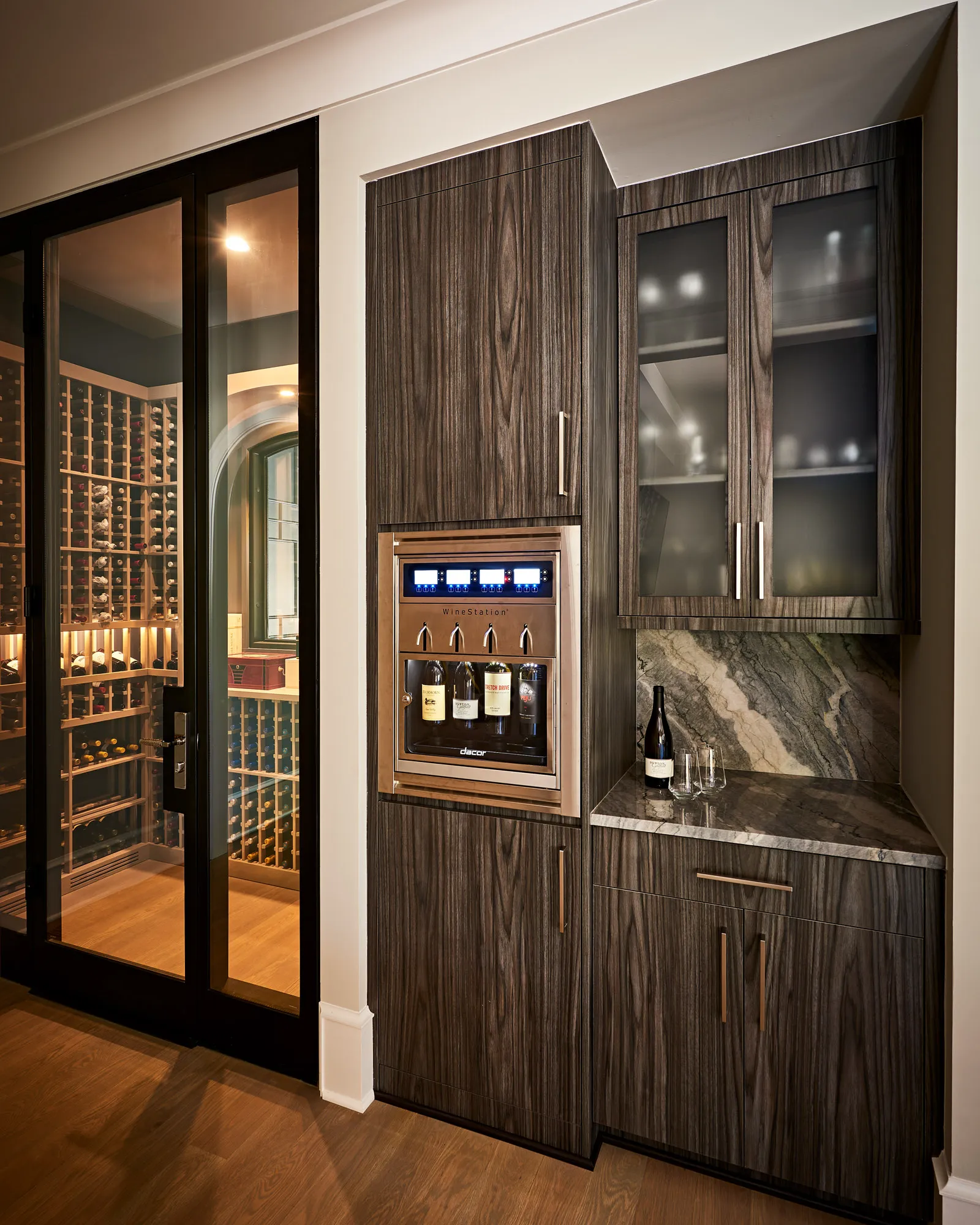 A modern, dark-grained wood wine station featuring a built-in dispenser and marble countertop. To the left is a sliding glass door opening to a large, illuminated wine cellar.