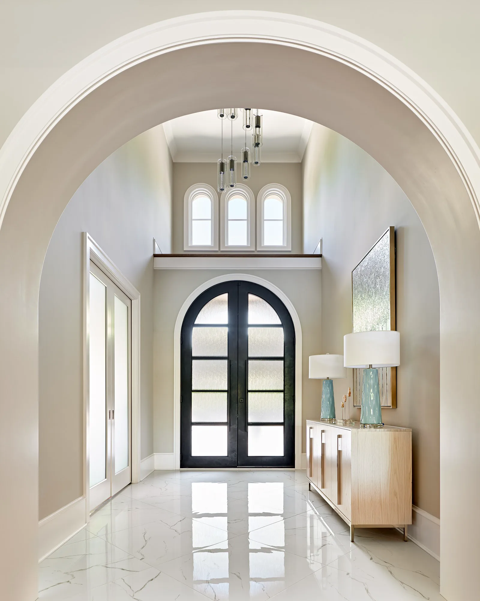 A modern, two-story foyer with polished marble floors, a black arched door, and a light wood console with two bright blue ceramic lamps.