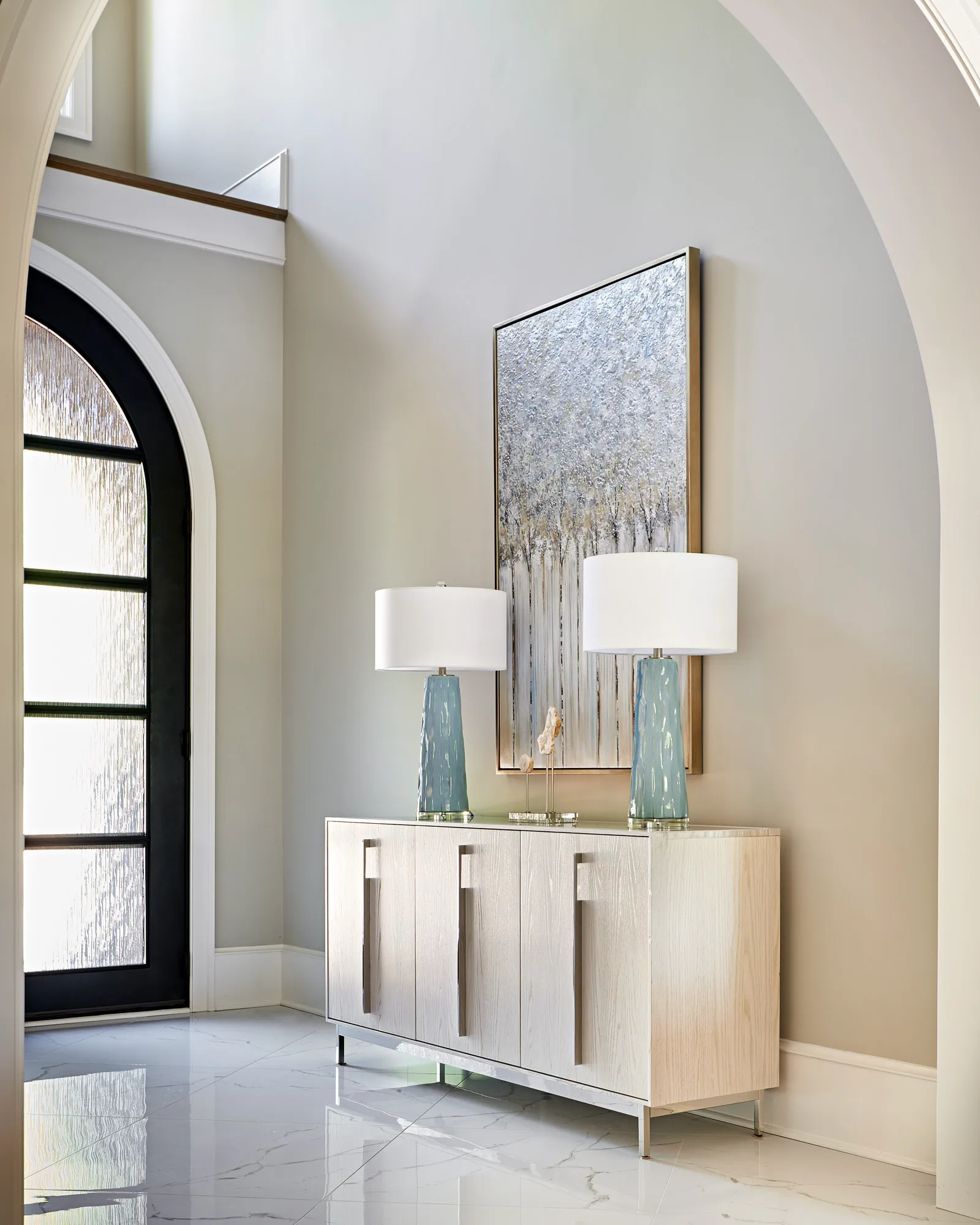 A modern foyer featuring a light wood cabinet topped with two matching blue glass lamps and an abstract silver tree painting, set against a polished marble floor.