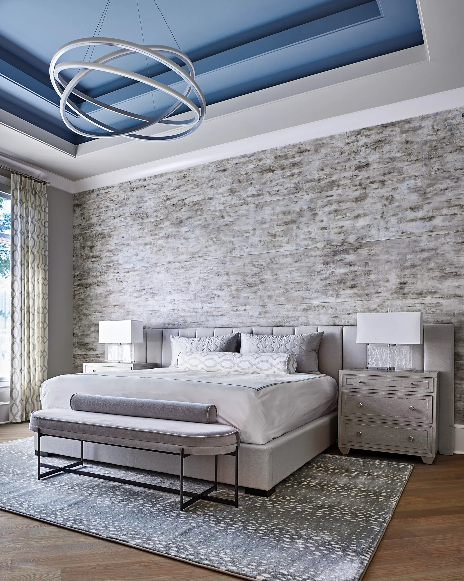 A stylish bedroom featuring a gray upholstered bed against a textured wall and a striking blue tray ceiling. A sculptural chandelier provides light over the space.