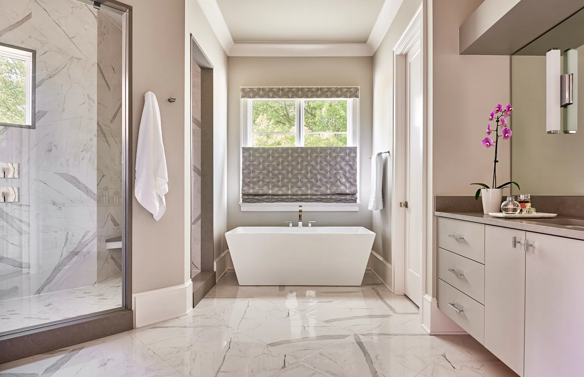 A luxurious, modern bathroom featuring glossy white marble flooring, a freestanding soaking tub centered under a window, a glass shower, and a clean gray vanity with an orchid.