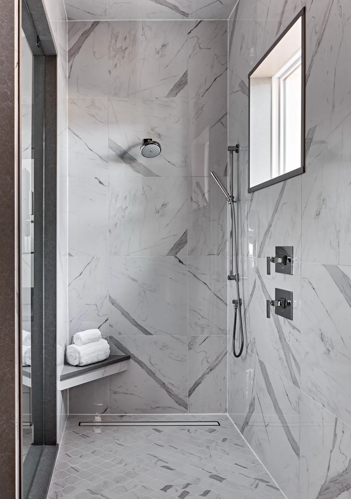 A sleek, modern walk-in shower with large white marble wall tiles and a gray mosaic floor. It includes chrome fixtures, a linear drain, and a bench holding rolled towels.