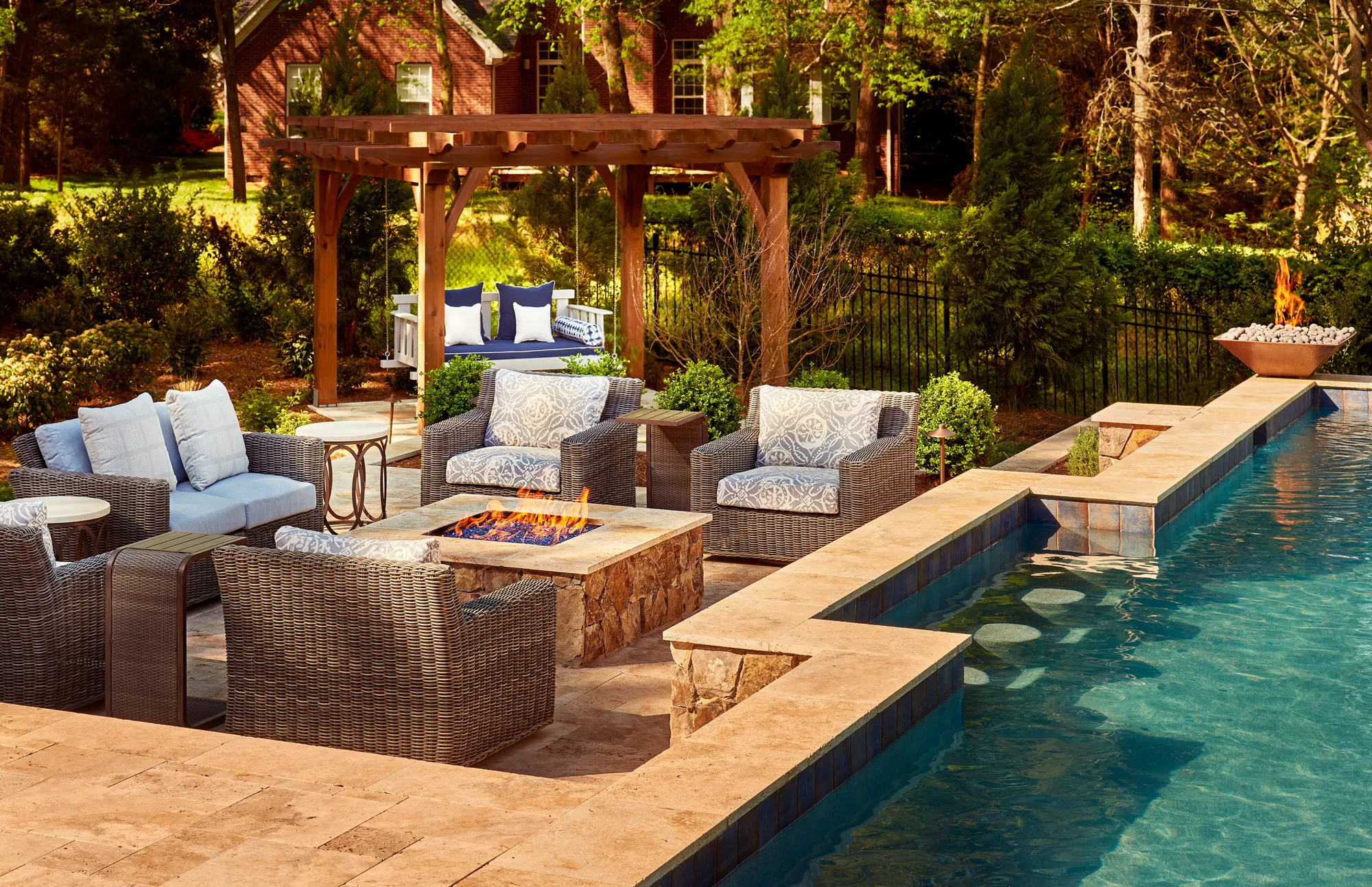 A luxurious outdoor patio next to a pool, featuring woven seating around a stone fire pit. A wooden pergola covers a swing seat in the lush green backyard.