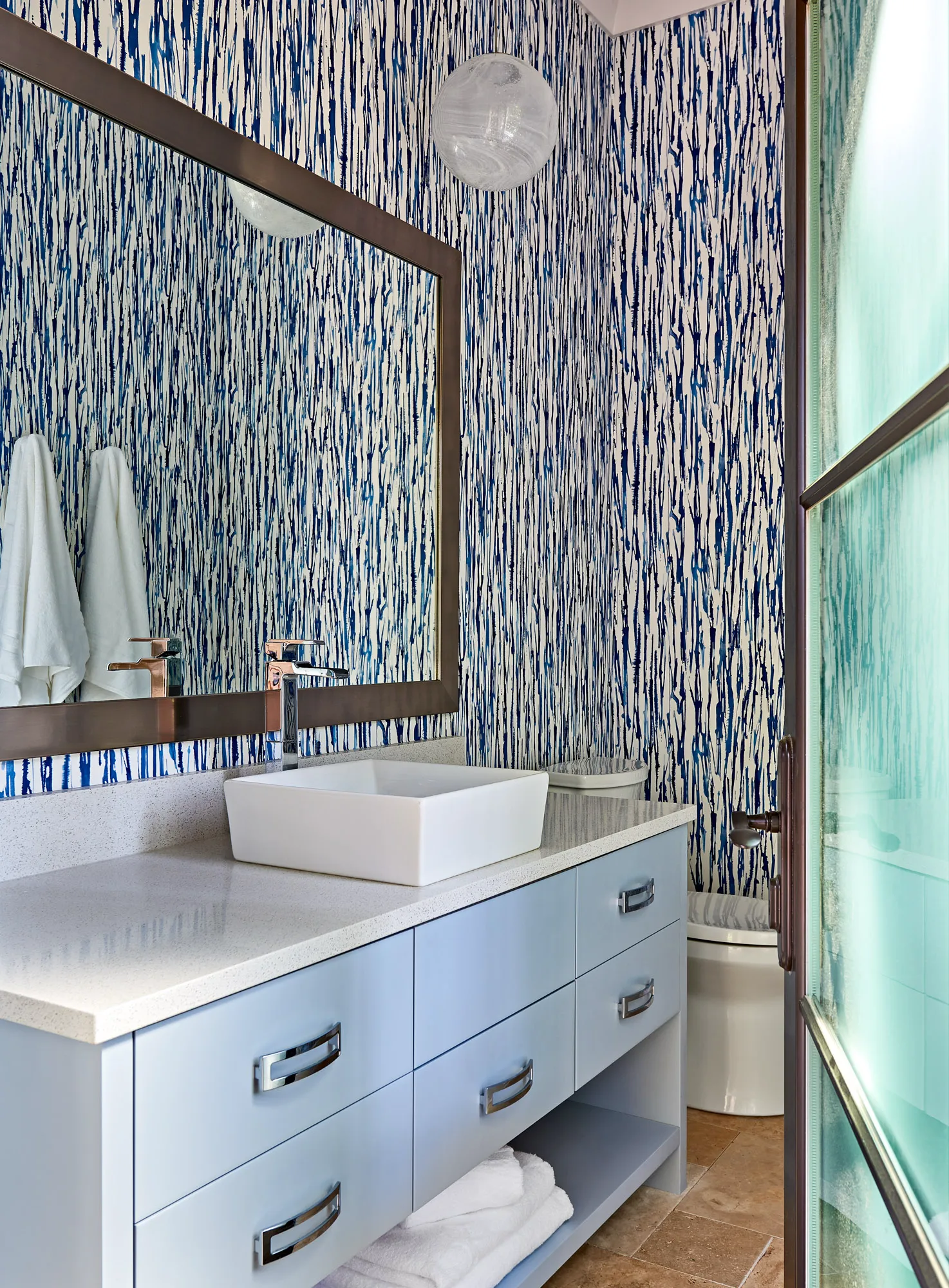 A contemporary bathroom with striking blue and white vertical wallpaper, a light blue vanity, white vessel sink, and chrome fixtures.