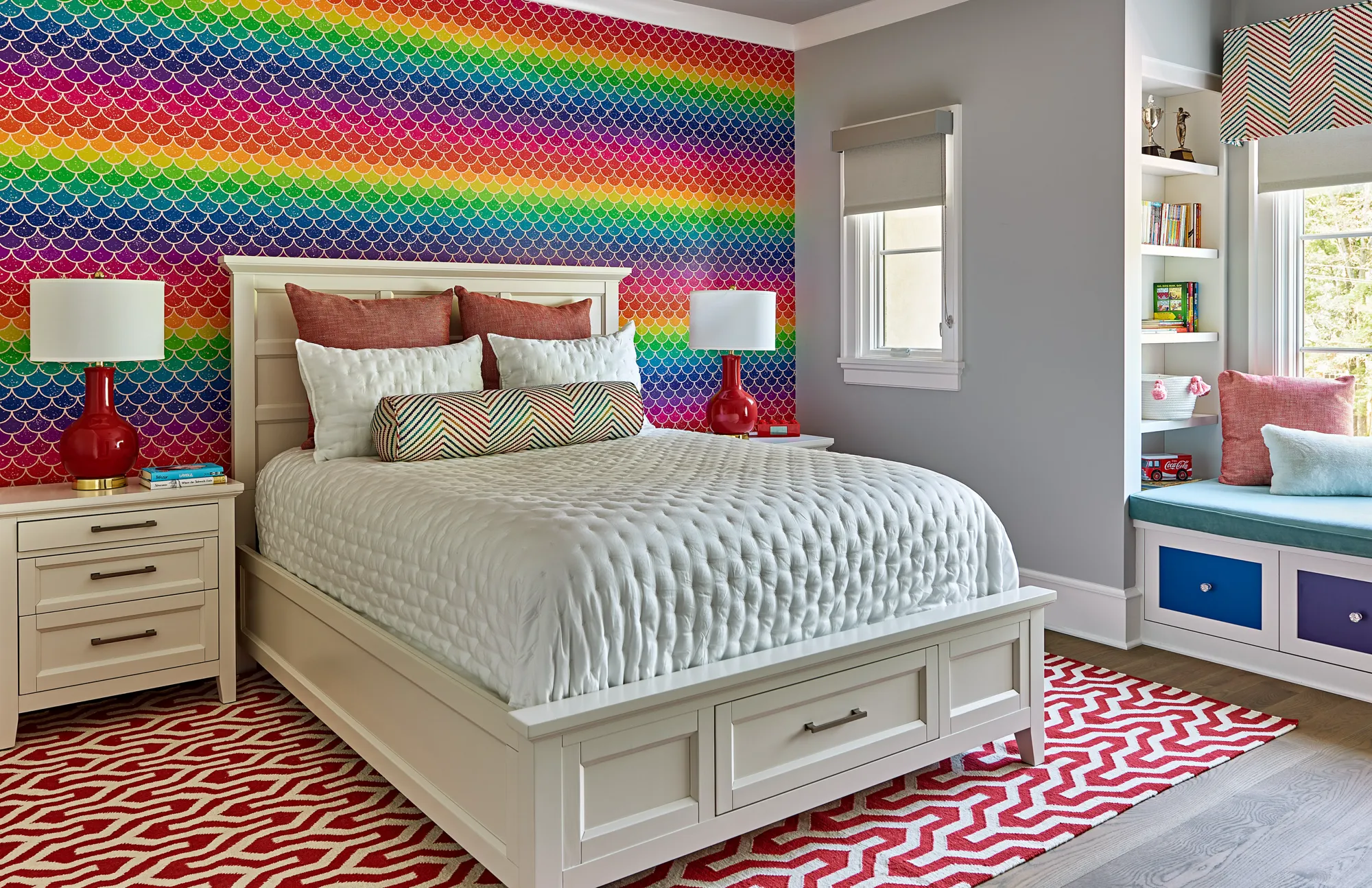 A playful bedroom featuring a white bed and furniture set against a vibrant rainbow, mermaid scale wallpaper. A red and white chevron rug covers the floor.