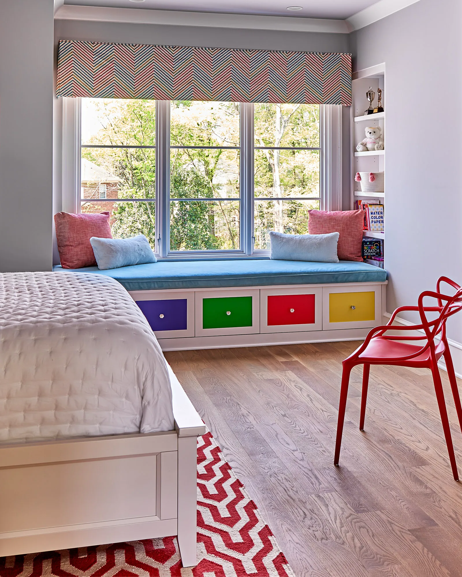 A child's room featuring a large window seat with a blue cushion and white built-in storage drawers painted in bright primary colors.