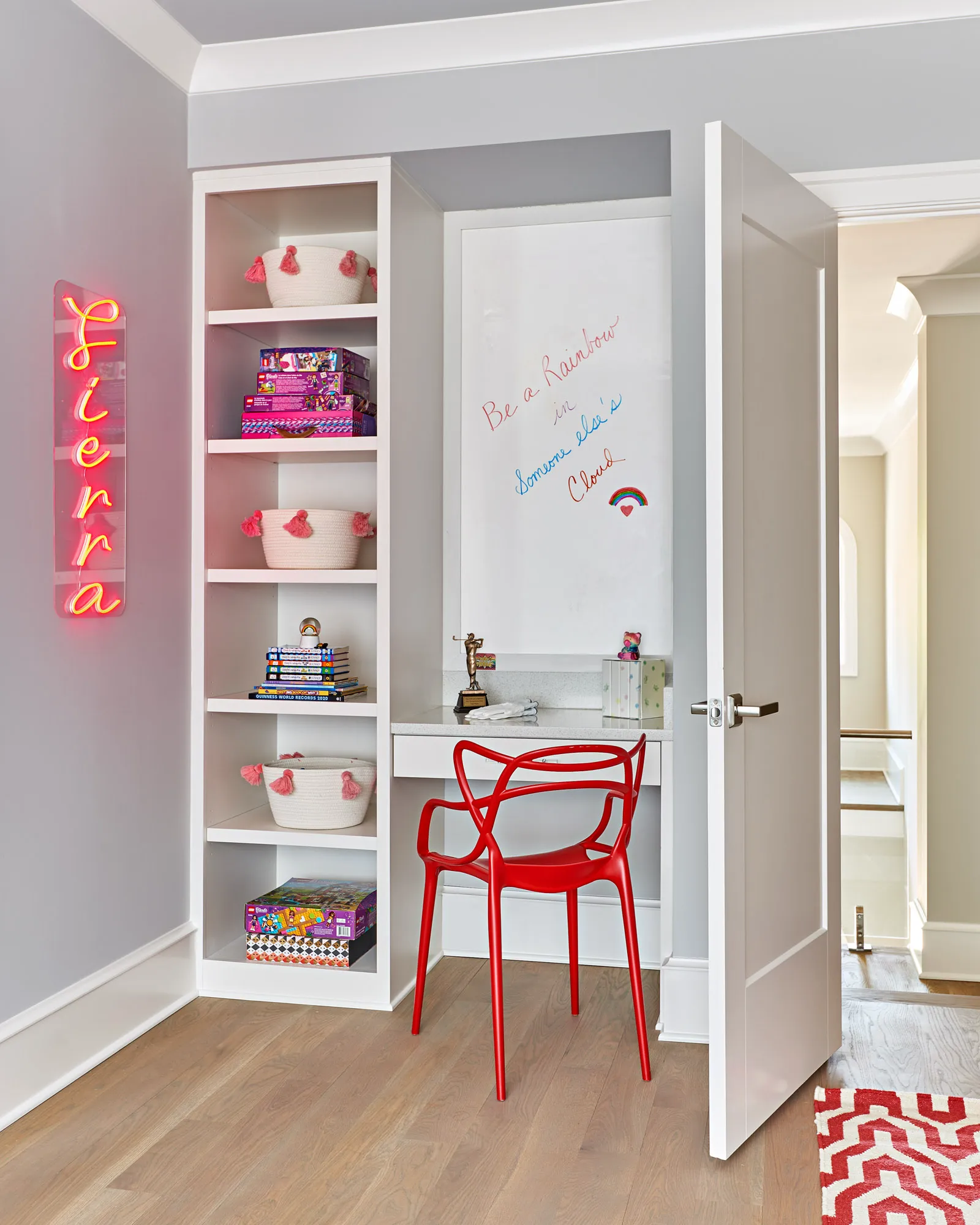 A built-in white desk nook features storage shelving, a bright red chair, and a whiteboard inscribed with "Be a Rainbow." The shelving holds playful baskets with pink tassels.