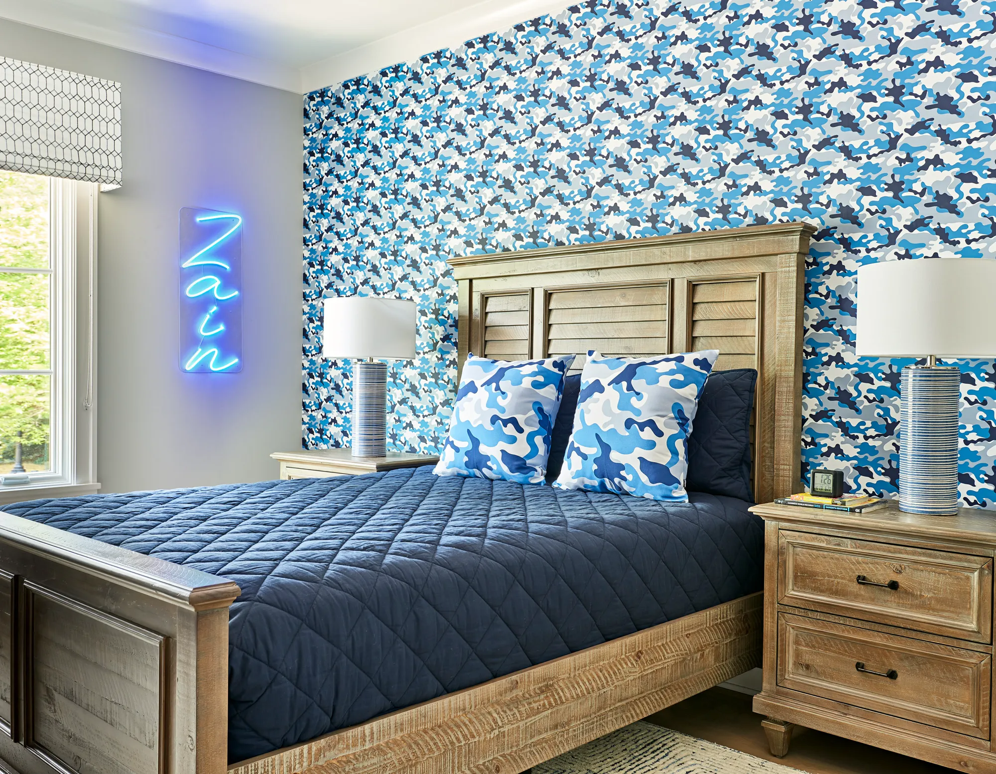 A rustic wooden bed with a navy comforter sits against a wall covered in blue and white camouflage wallpaper. The room includes matching nightstands, cylindrical lamps, and a personalized blue neon sign displaying the name "Zain."