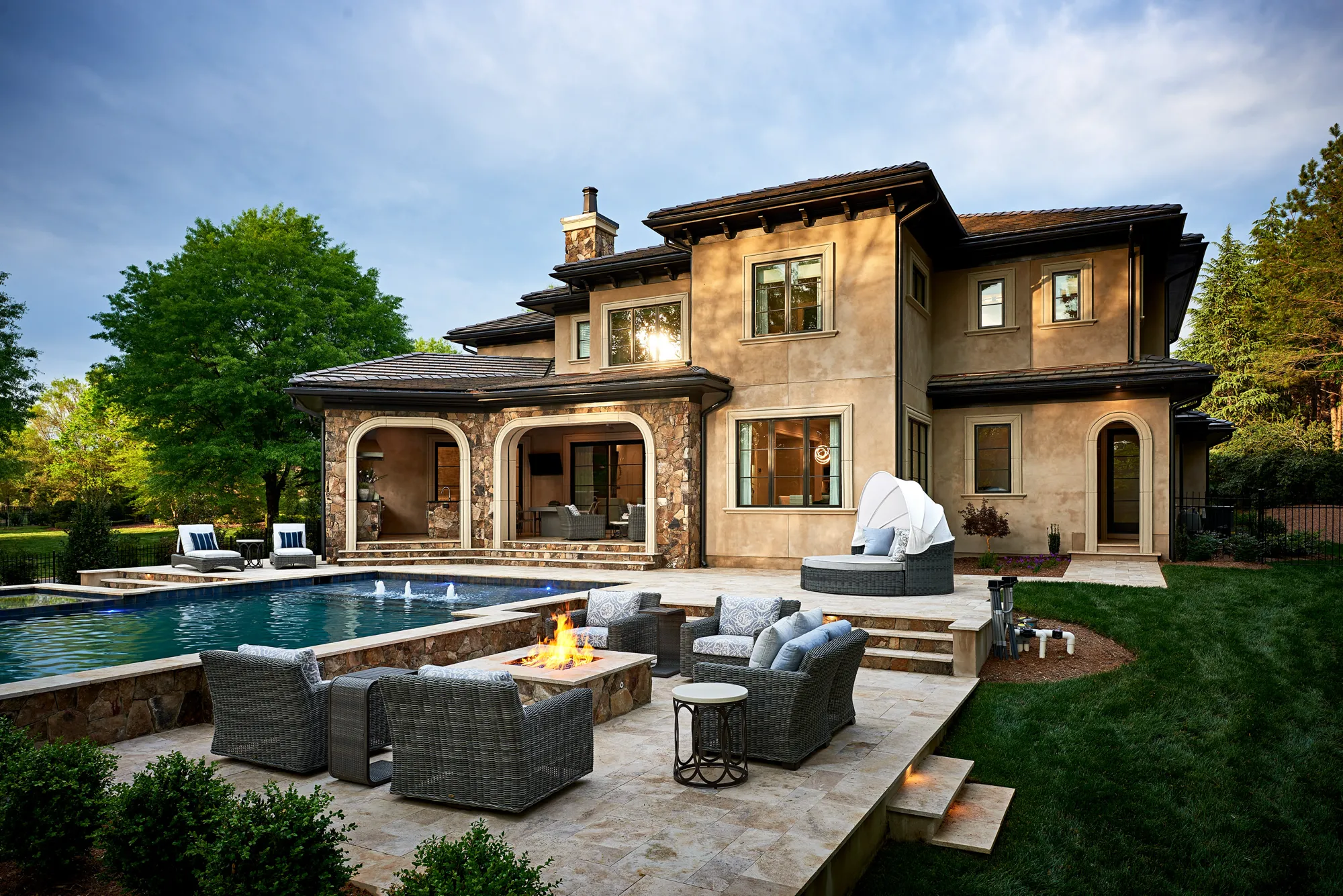 The exterior of a large stucco home with a travertine patio designed for outdoor living. The space features a lit fire pit, woven seating, and a rectangular swimming pool.