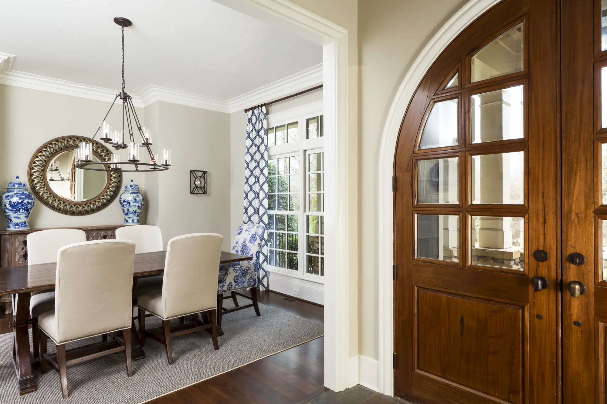 View into an elegant dining room with a dark wood table and upholstered chairs, framed by a decorative arched wooden entryway door on the right.