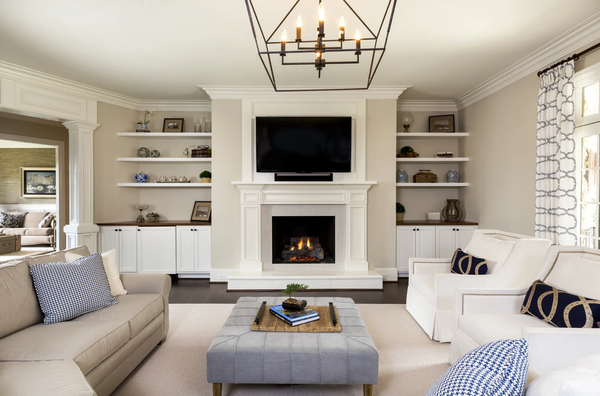 A traditional living room featuring a white fireplace with built-in shelves and a mounted TV. The neutral furniture includes a beige sofa, white chairs, and a gray tufted ottoman.