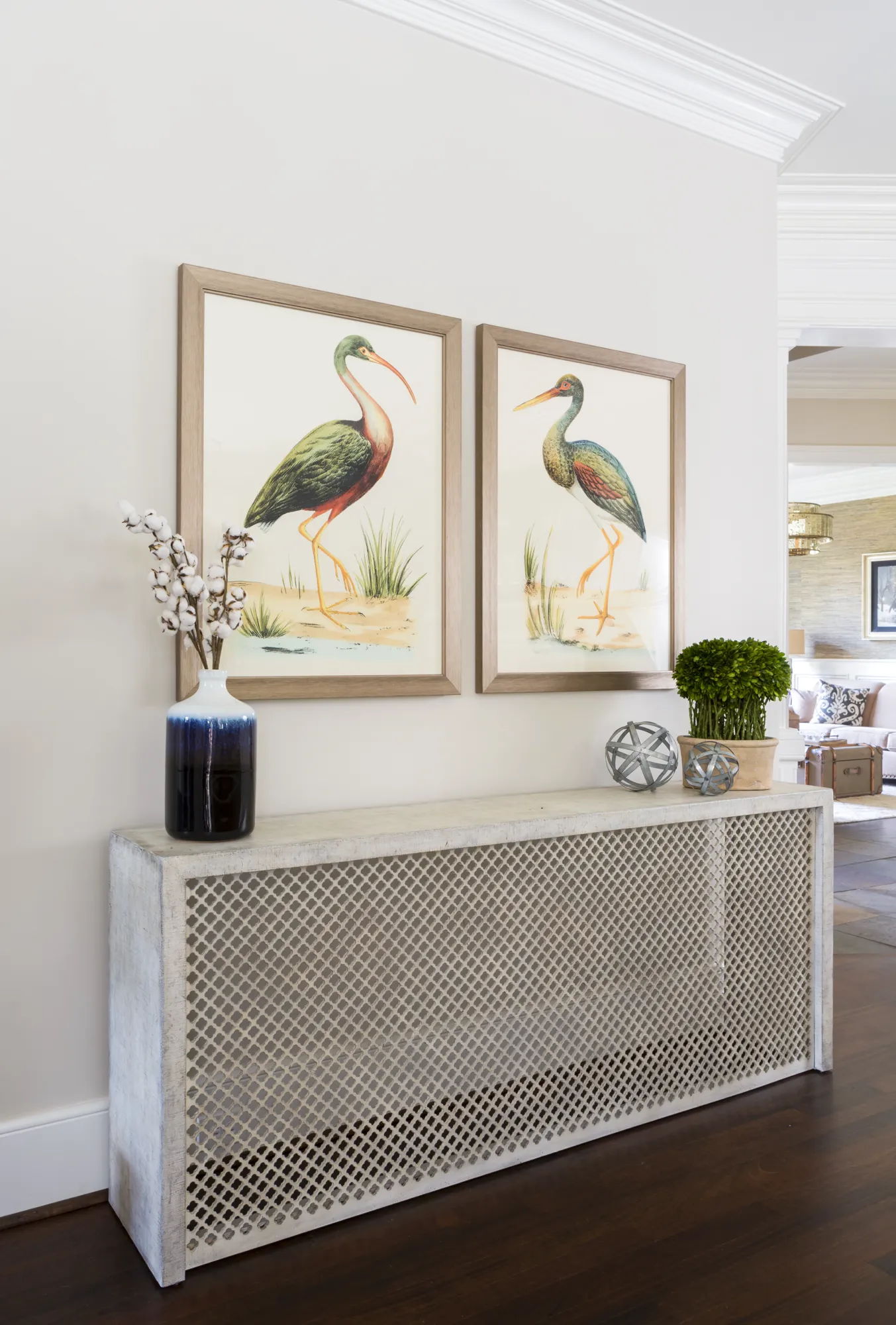 A lattice-front console table beneath two framed prints of colorful wading birds. The table is decorated with a vase holding cotton branches and a potted green plant.