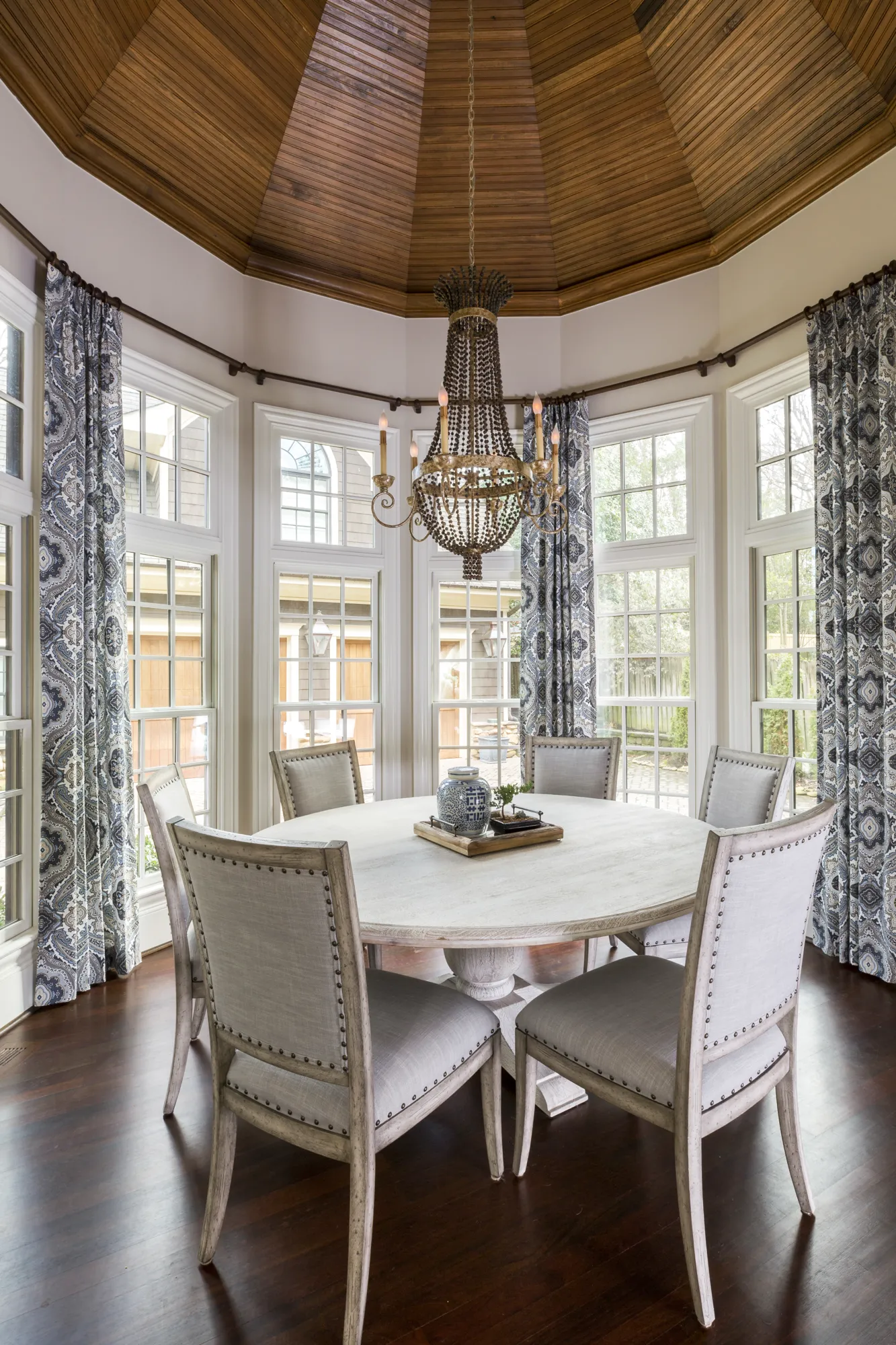 A bright dining area features a round table surrounded by six linen chairs on dark wood floors. The room is enclosed by many windows with blue patterned drapes, centered beneath a dark wood vaulted ceiling and a beaded chandelier.