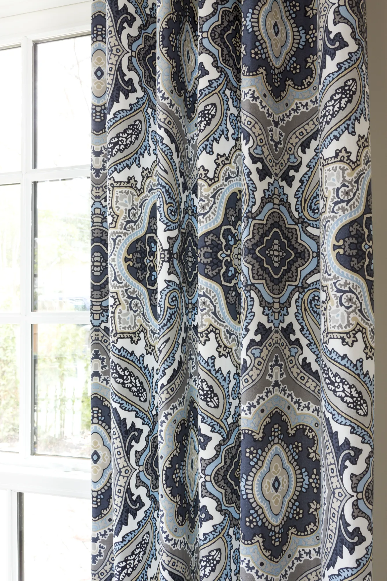 A patterned curtain with large folds featuring a damask design in navy, light blue, gray, and white, positioned next to a window.