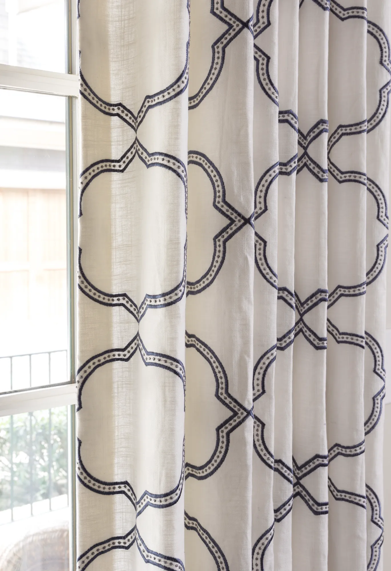 A pleated white linen-look curtain with an embroidered navy blue geometric trellis pattern hangs next to a sunlit window.