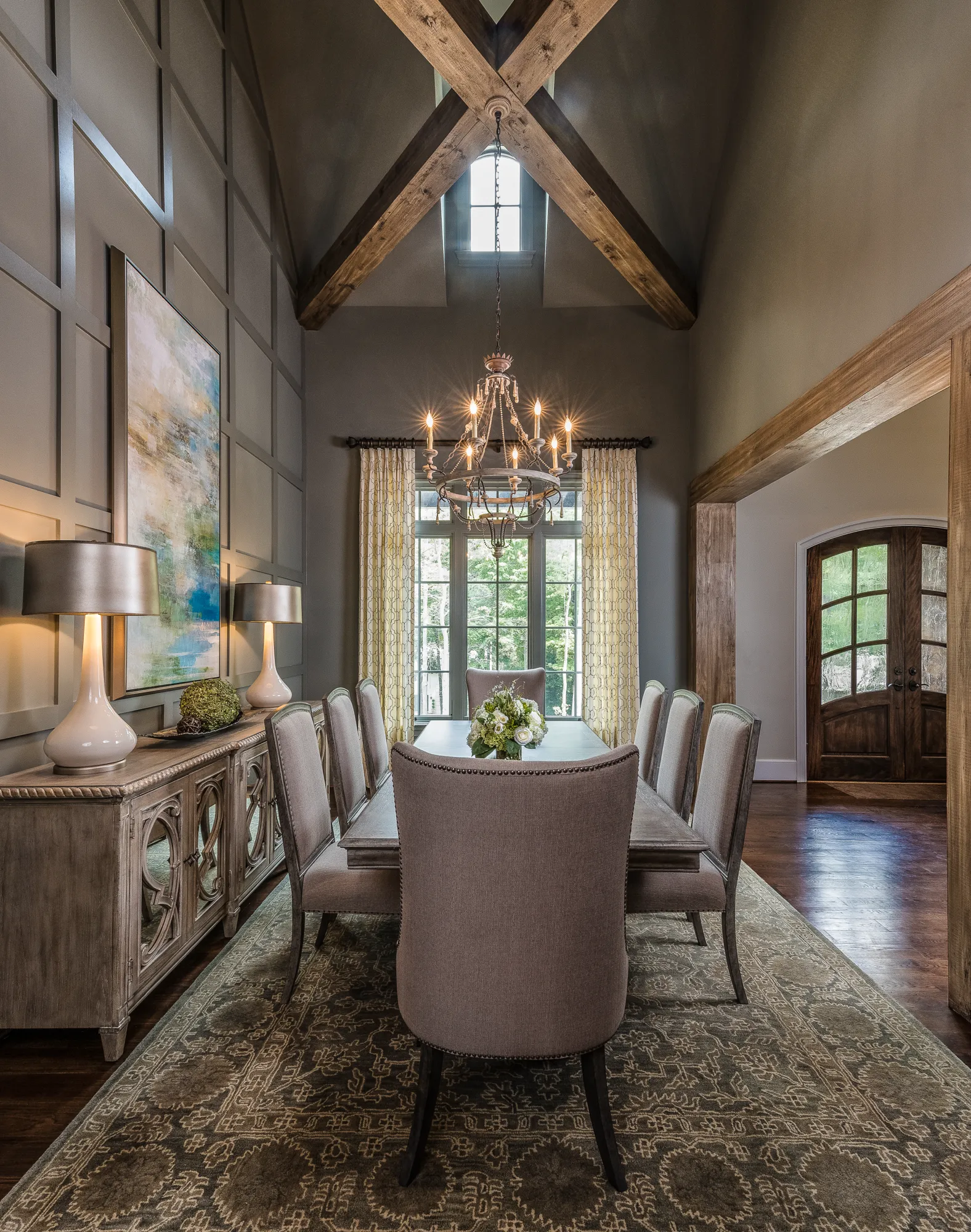 A sophisticated dining room with a high, beamed ceiling, large chandelier, and a long wooden table surrounded by taupe upholstered chairs. Dark paneled walls and a patterned rug complete the look.