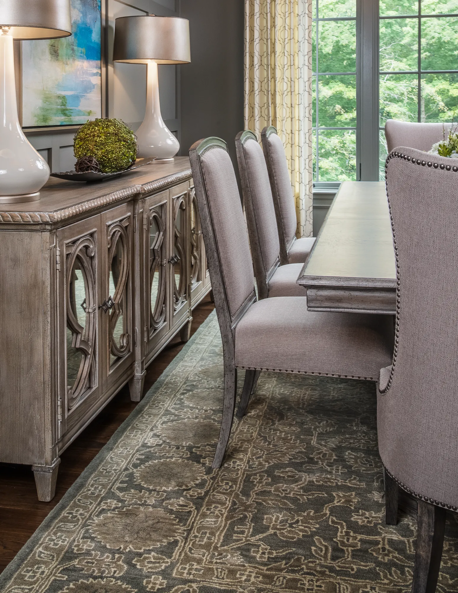 A formal dining space with a distressed wood credenza and two tall white lamps, set against a dark gray wall. Upholstered dining chairs line the table next to a window view.