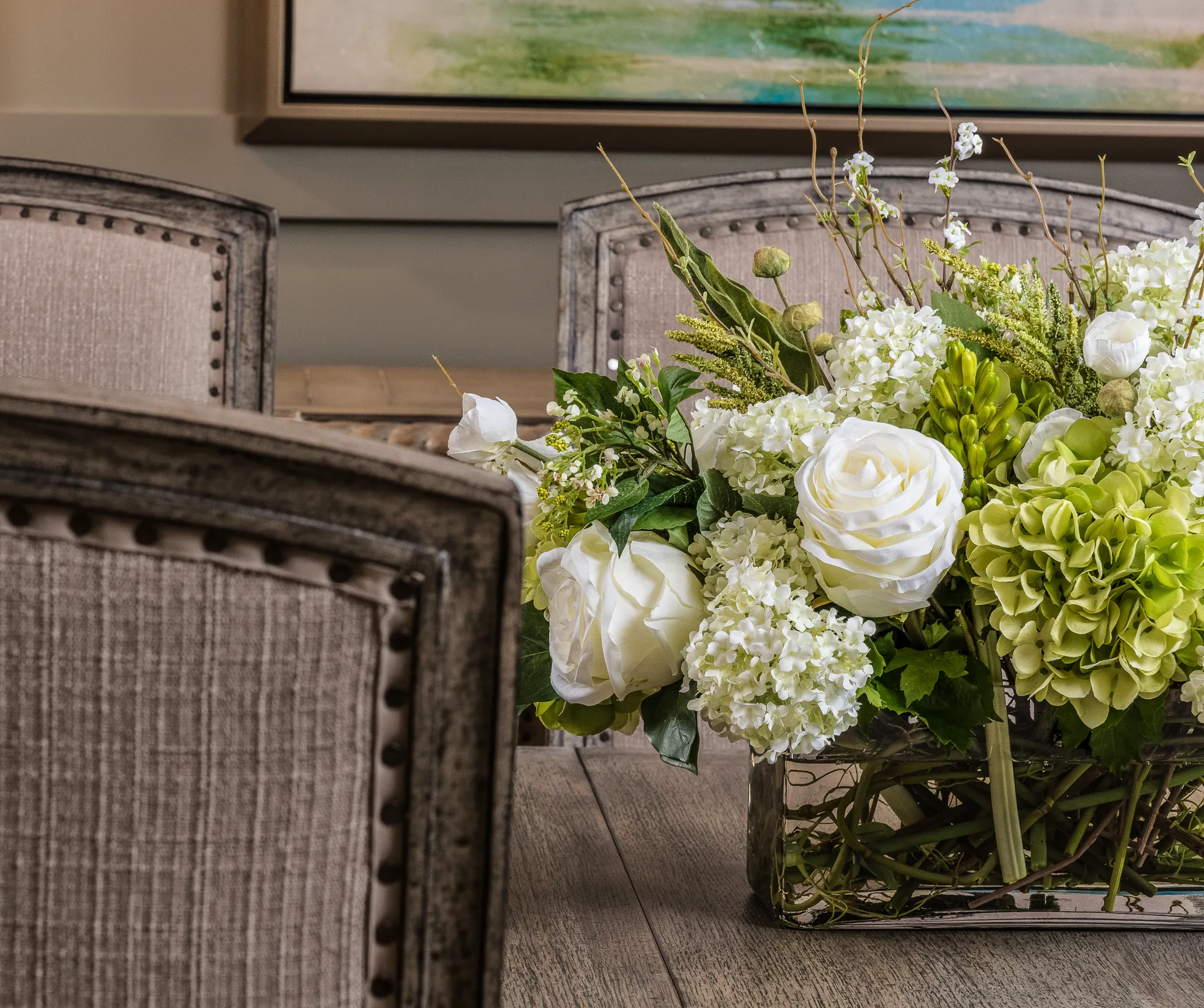 A large arrangement of white roses and green hydrangeas in a glass vase is centered on a wooden table, framed by the backs of upholstered dining chairs.