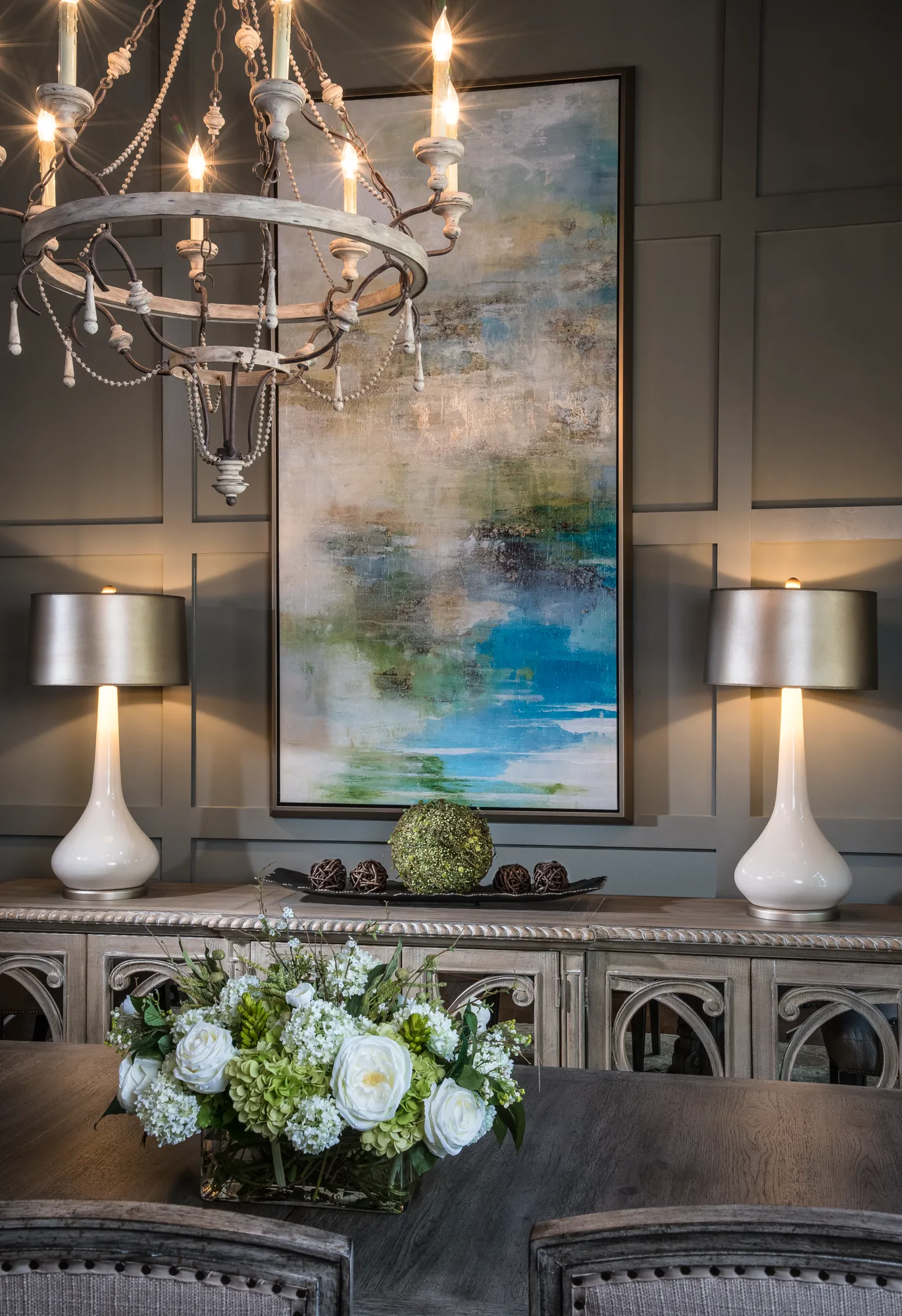 A distressed wood buffet table against a paneled gray wall features two white lamps and an abstract painting. A rustic chandelier hangs above a foreground bouquet of white and green flowers.