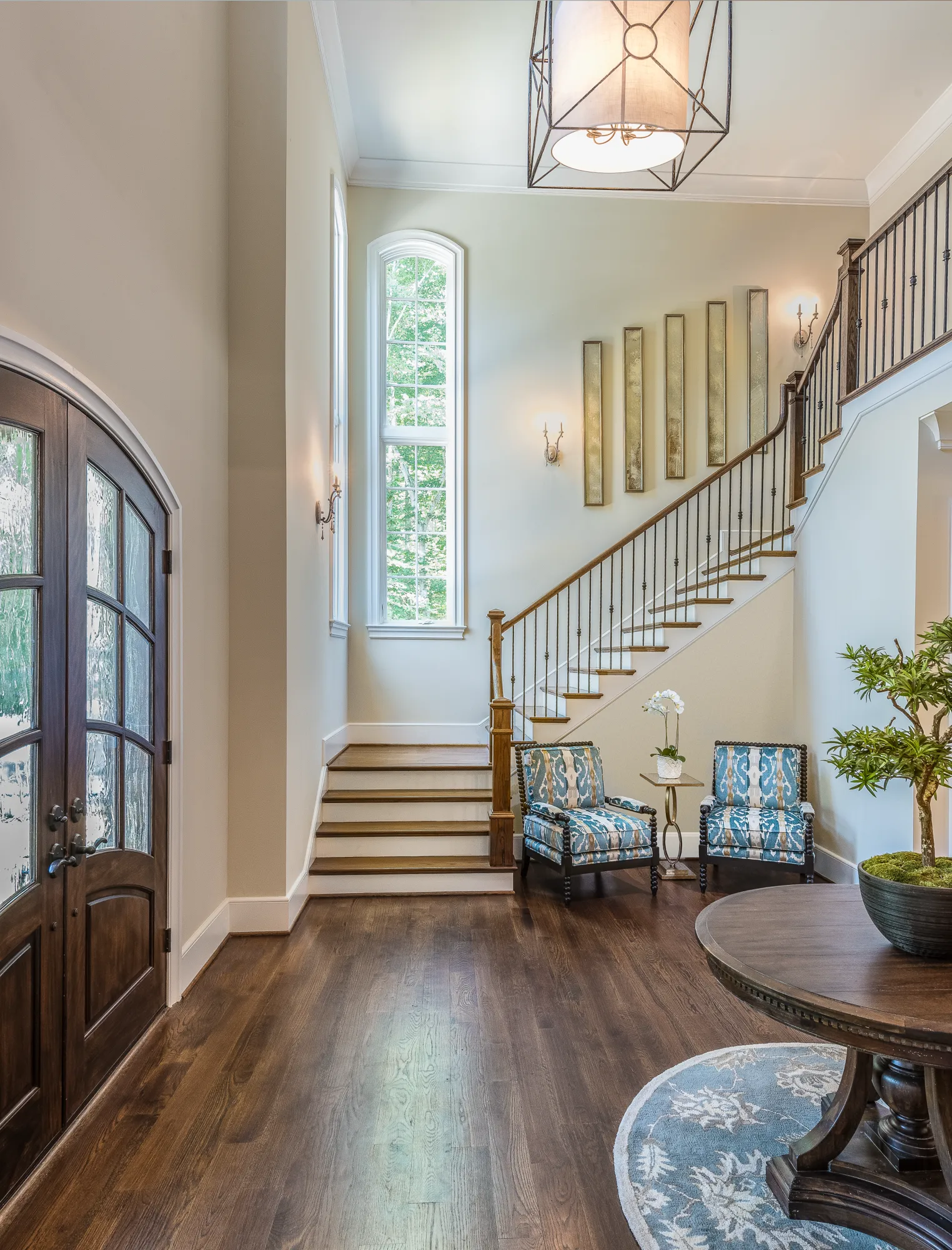 An elegant two-story foyer with dark wood floors, double arched entry doors, a sweeping staircase, and a sitting area featuring two patterned chairs.