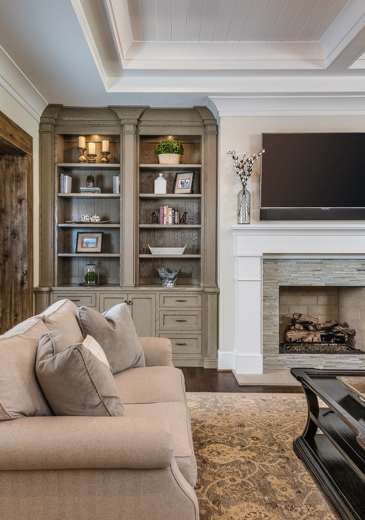A transitional living room with a neutral sofa, a stone fireplace, and custom built-in bookshelves painted a gray-washed wood tone.