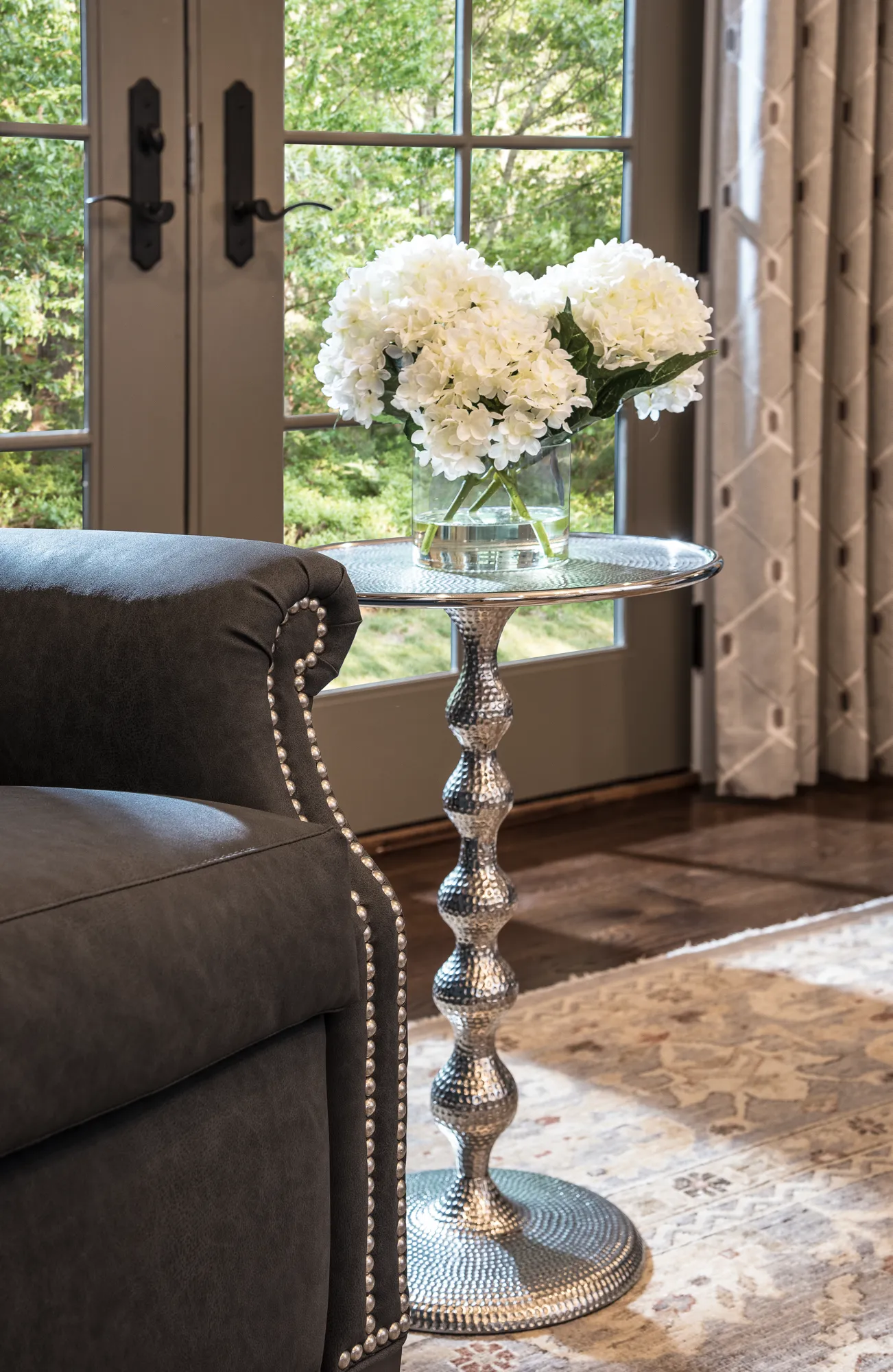 A dark gray armchair with silver nailhead trim next to a hammered silver pedestal side table holding white hydrangeas.