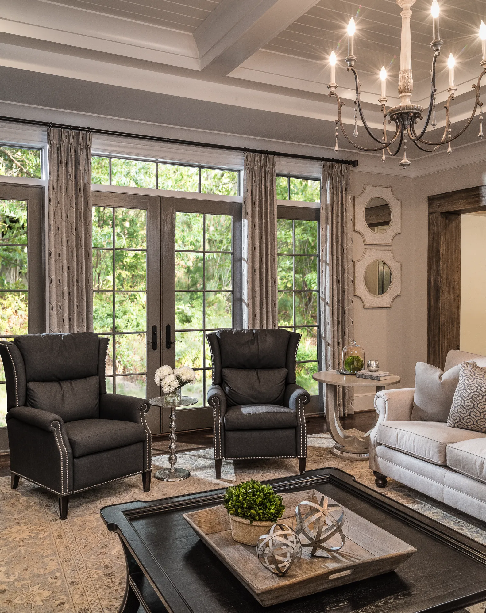 Two dark gray wingback chairs face a light sofa in an elegant living room. A decorative chandelier hangs from the coffered ceiling above the furniture grouping.