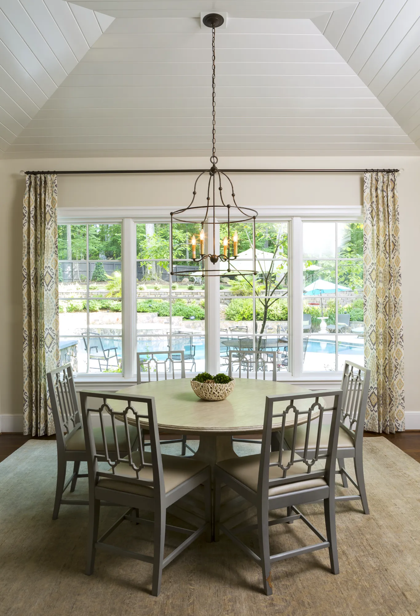 A round dining table surrounded by six gray geometric-back chairs sits under a vaulted ceiling. Large windows reveal a lush backyard and swimming pool.