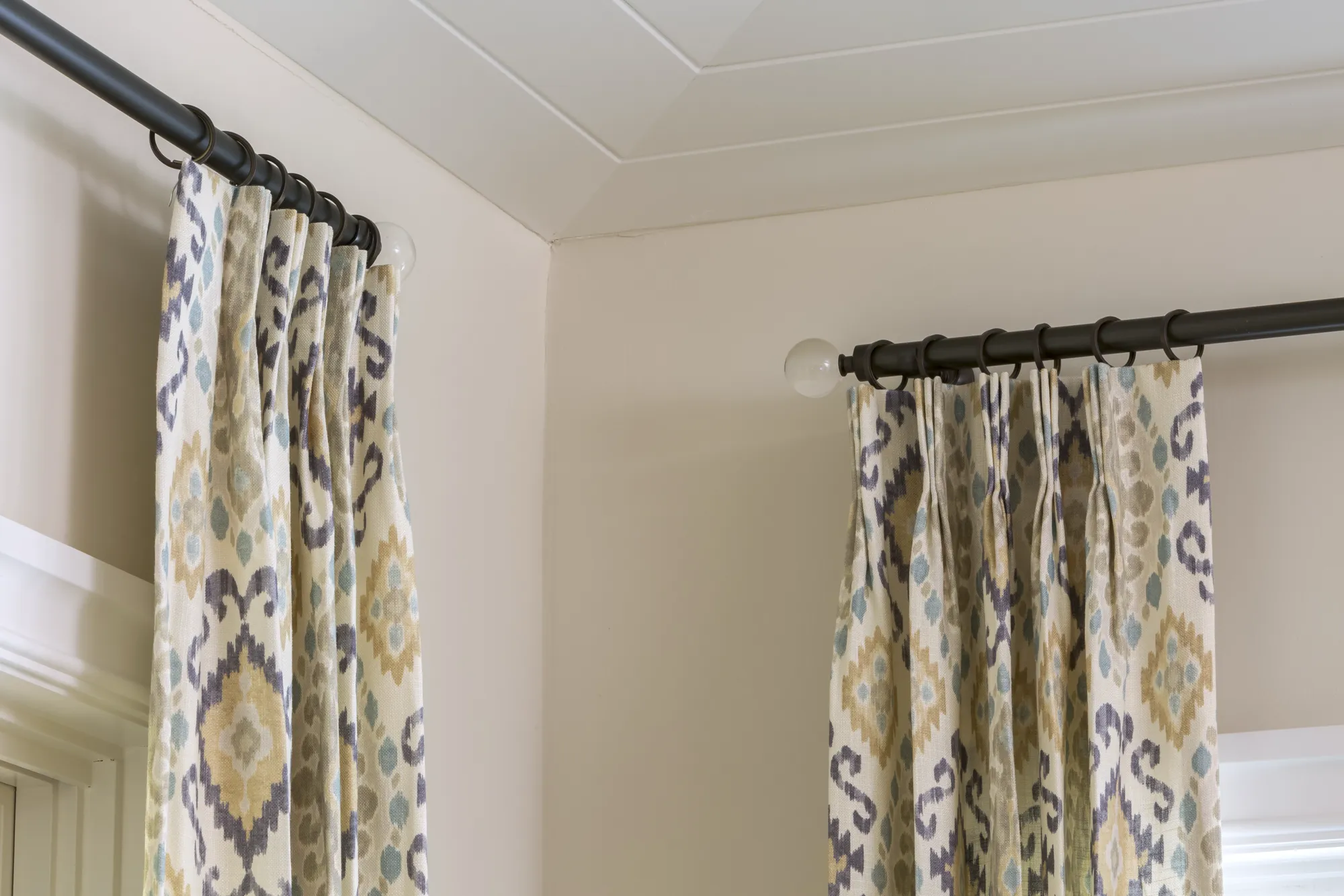 Close-up of two pleated window curtains featuring a colorful ikat pattern in beige, blue, and purple, hung on a dark rod with clear finials near a corner.