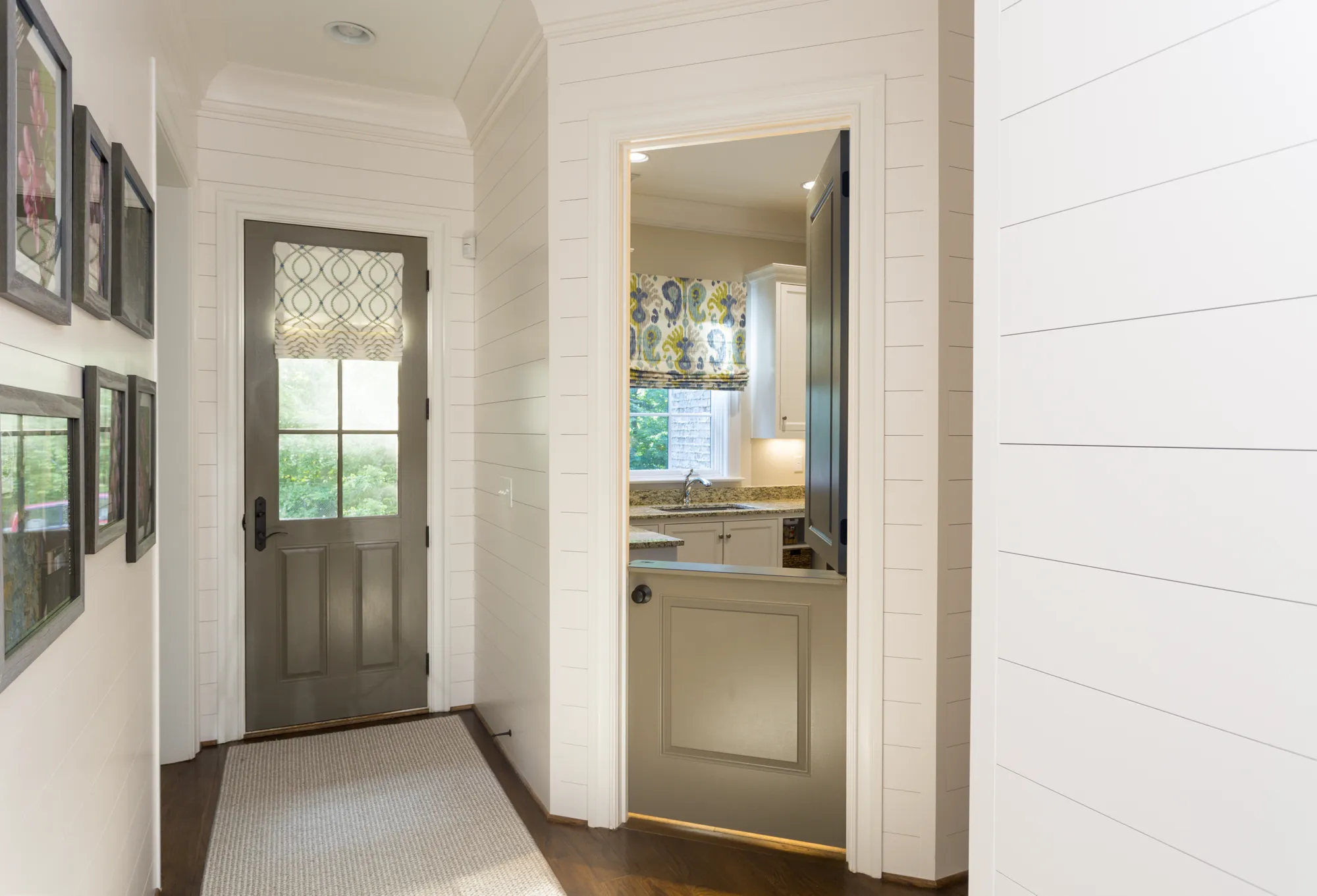 A white shiplap hallway features a gray exterior door with a patterned shade and a runner rug. A half-door opening leads to a room with a sink and white cabinets.