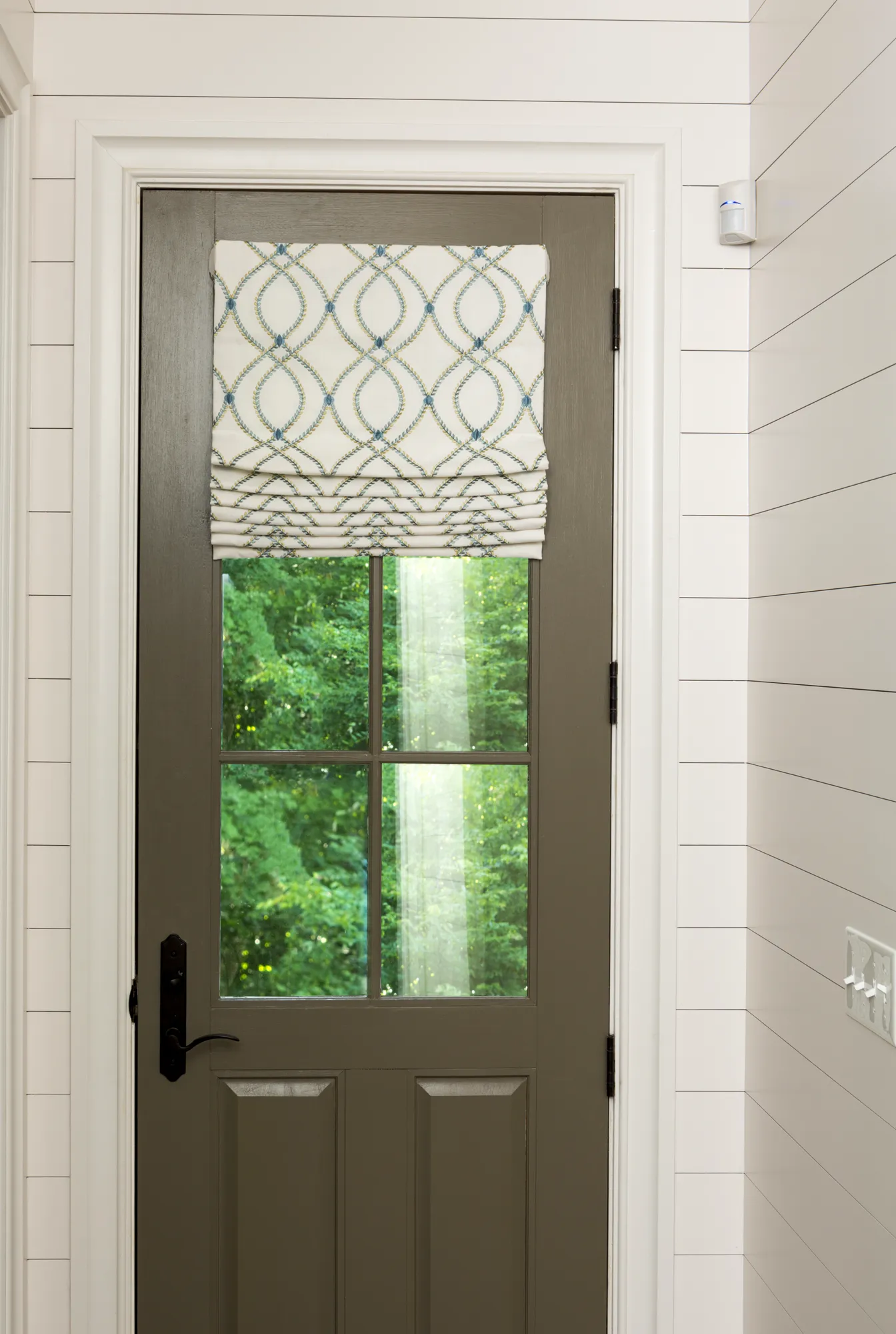 A deep olive-brown exterior door, surrounded by white shiplap walls, features a patterned Roman shade pulled down over the glass pane that overlooks green trees.