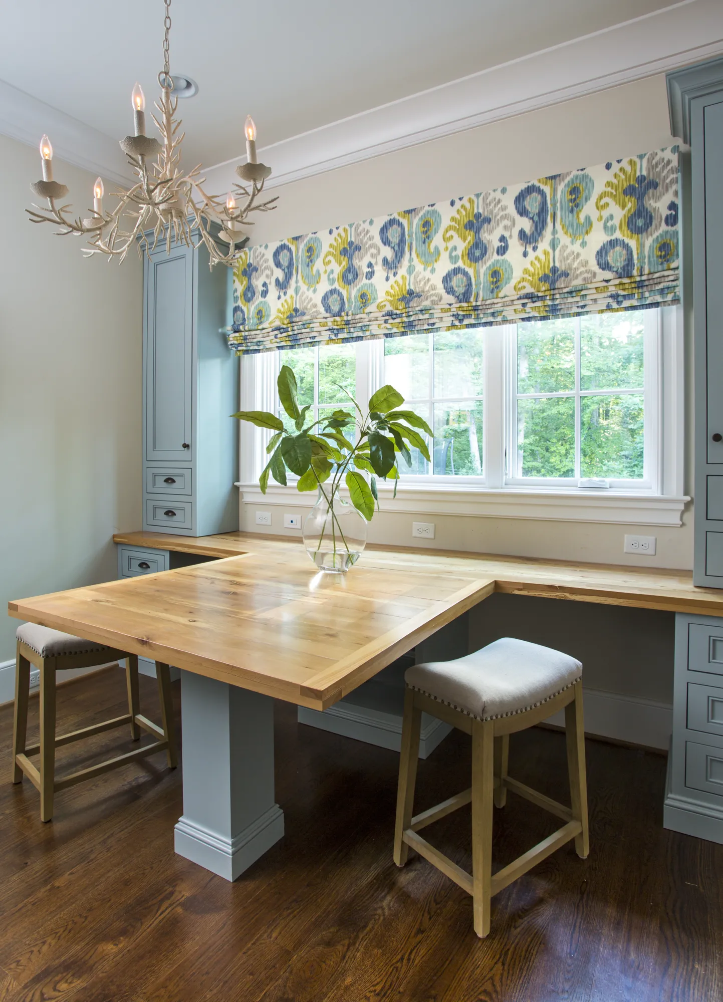 A custom home office nook with blue built-in cabinets and a light wood desk. The window has a blue and yellow patterned Roman shade.
