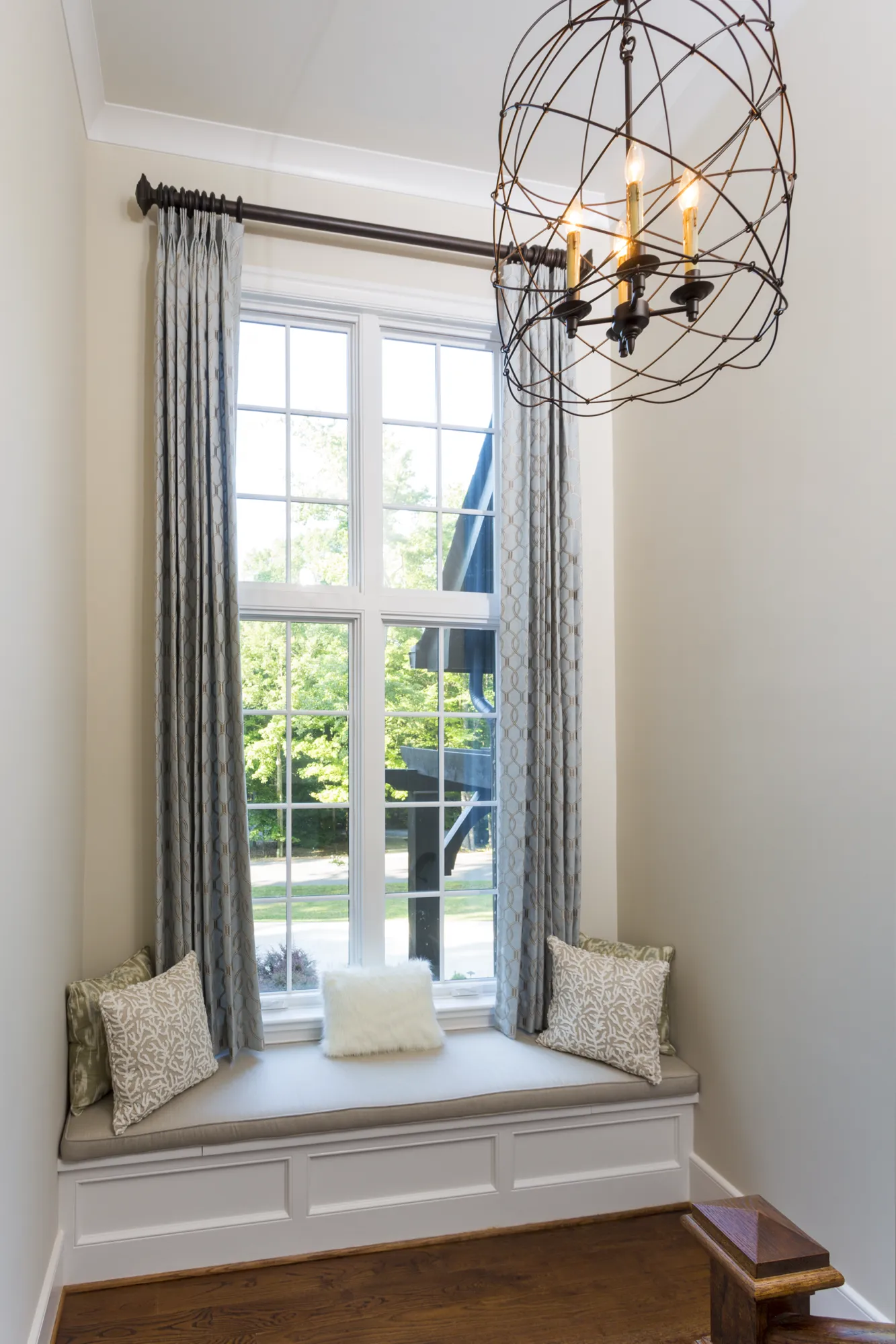 A tall, light-filled window features a cushioned window seat decorated with throw pillows. The area is illuminated by a geometric dark metal orb chandelier hanging above patterned blue drapes.