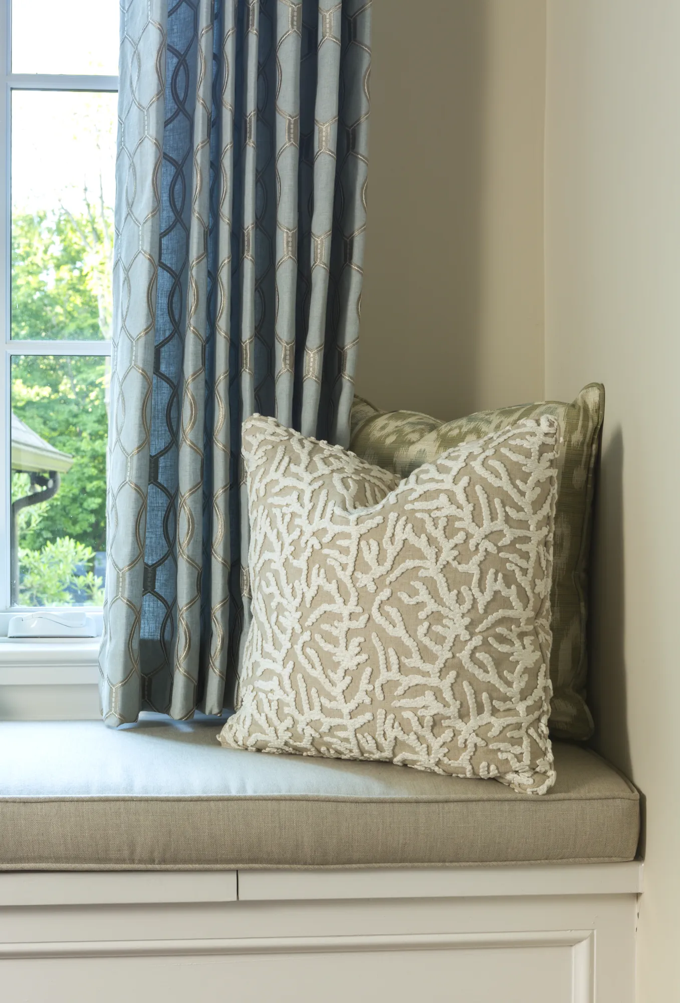 A cushioned window seat with custom blue geometric drapery and two decorative throw pillows, one with white coral embroidery.
