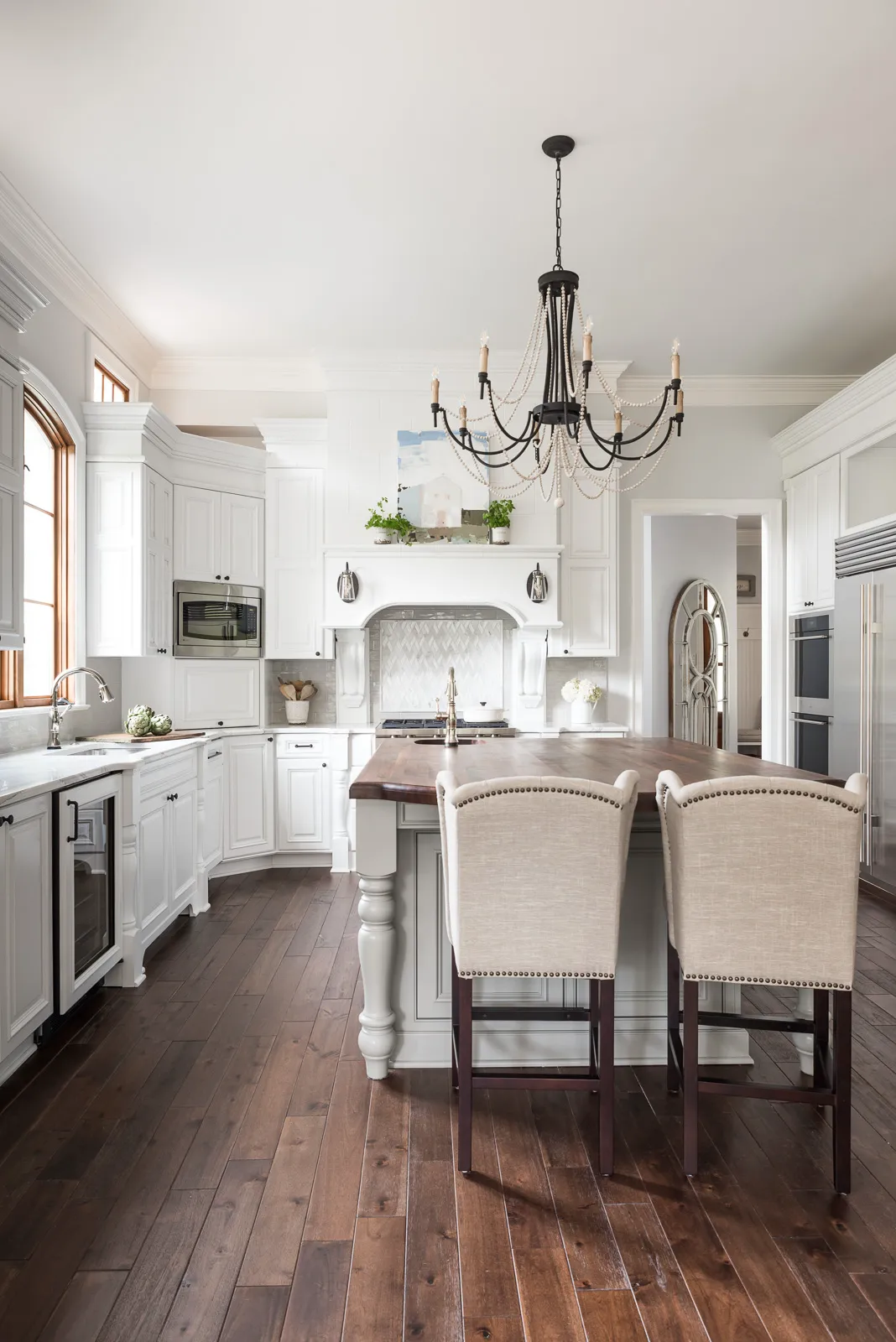 A white, high-end kitchen with dark hardwood floors, featuring white perimeter cabinetry and a large central island with a wooden countertop and two upholstered stools.