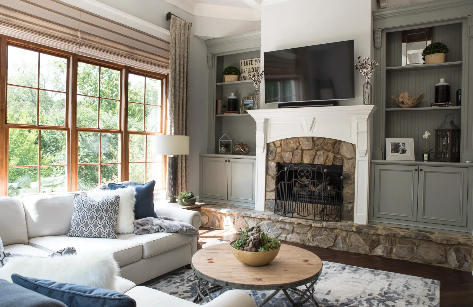 A light-filled living room features a white sofa and a stone fireplace flanked by gray built-in shelves. A wooden coffee table sits on an area rug near large windows.