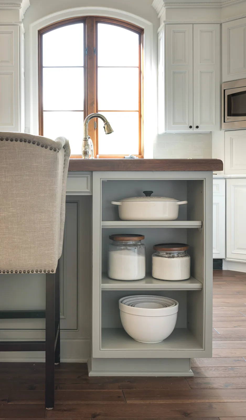 A light gray kitchen island with a wood counter has open shelving displaying white jars of ingredients and mixing bowls. A brass faucet is visible against a bright arched window.