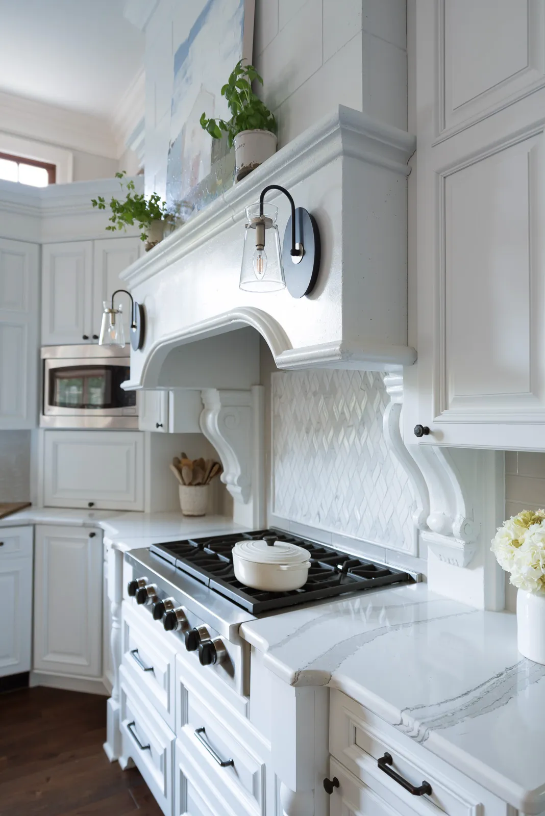 A bright white kitchen highlighting a stainless steel gas range and a large, detailed custom range hood with flanking glass and black wall sconces.