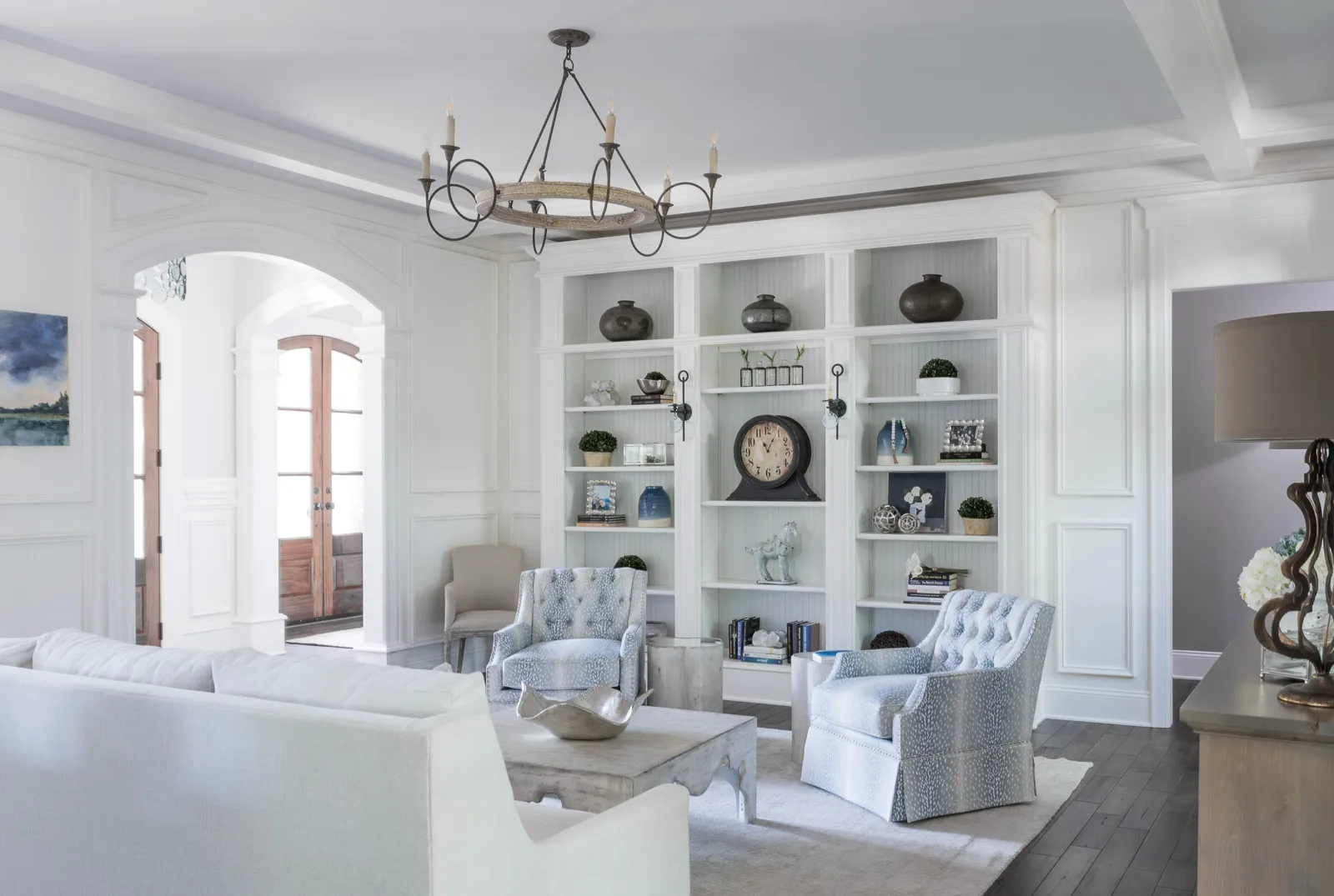 An elegant living room featuring white wainscoting and built-in bookshelves filled with decorative objects. Two blue patterned tufted armchairs and a white sofa furnish the space.