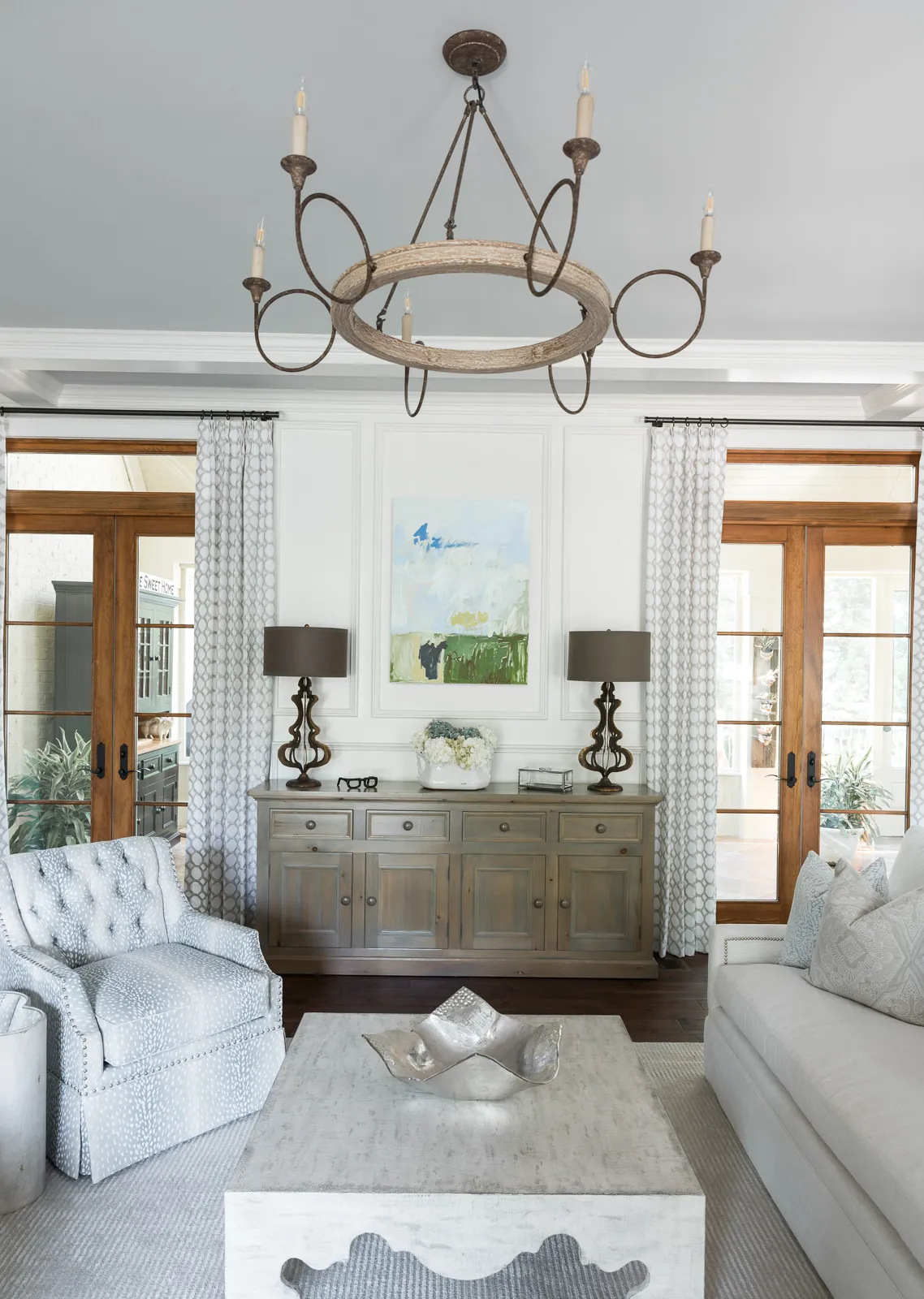 A bright, elegant living room with a rustic chandelier over a square coffee table, light-toned seating, and a centered credenza against a paneled wall.