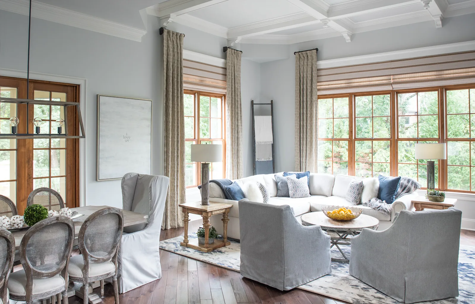 A tranquil open-concept living area with a white sectional, gray seating, and large wood-framed windows next to a rustic dining table.