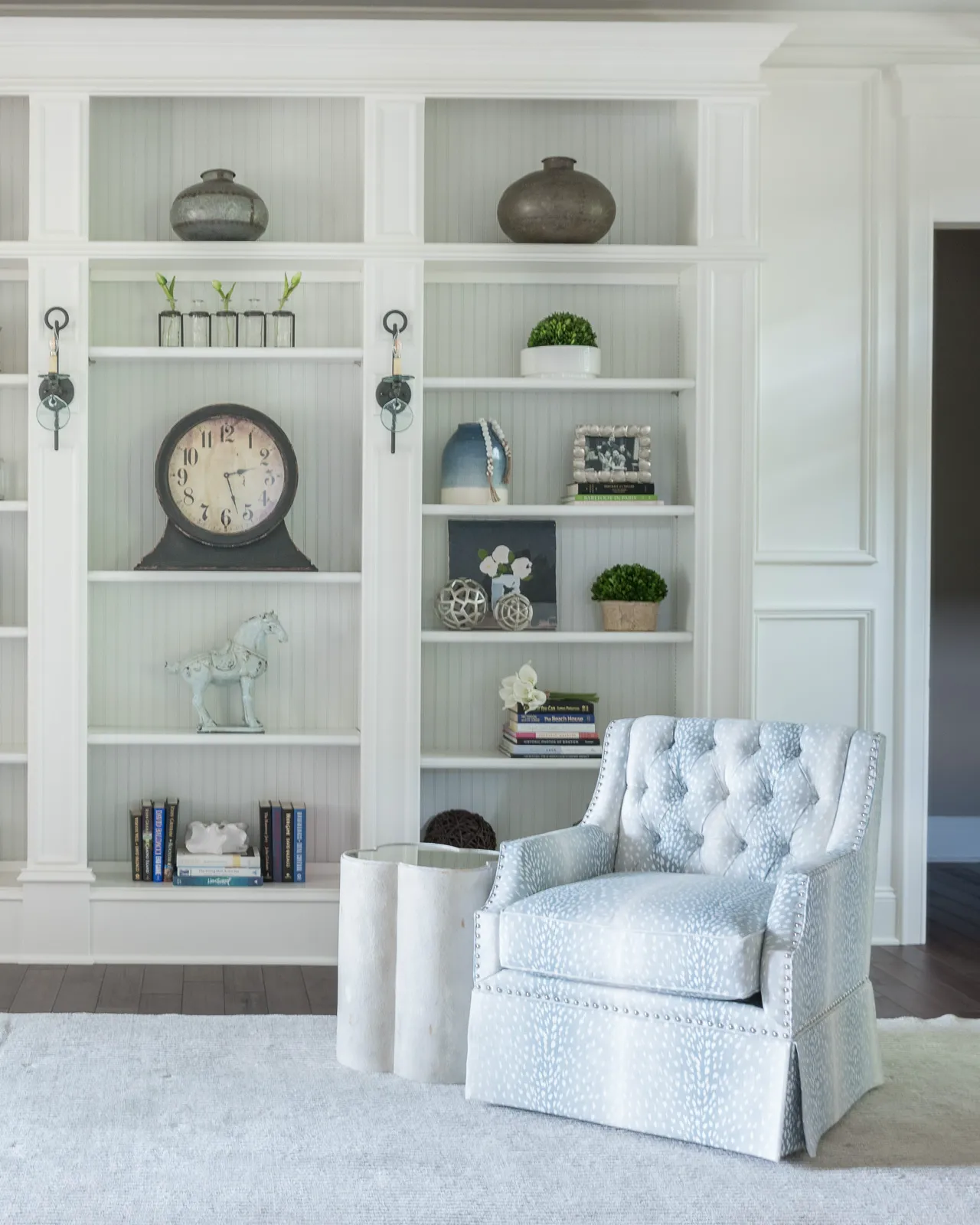 A tufted armchair in a light blue and white spotted fabric rests on a gray rug, positioned before elegant white built-in shelves displaying decor and books.