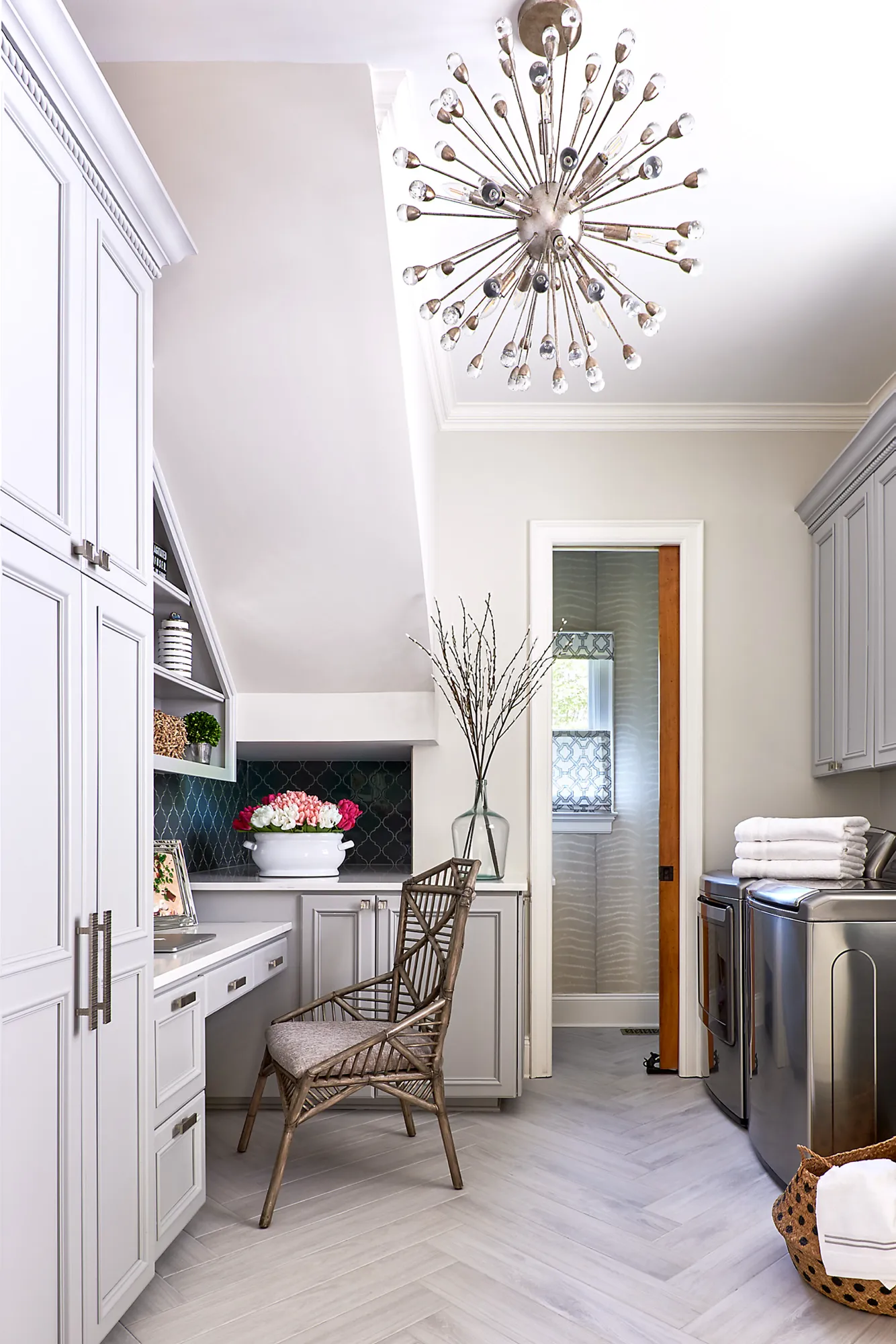 A well-lit, luxury laundry room and office space combo. It features light gray cabinets, a rattan chair at a built-in desk, and a unique crystal light fixture.