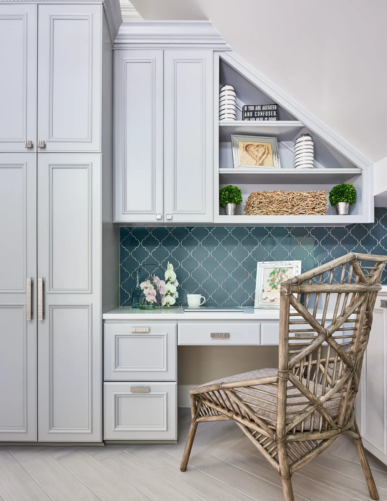 A built-in desk with light gray cabinetry and a teal tiled backsplash. A woven rattan chair is positioned at the counter, decorated with flowers.